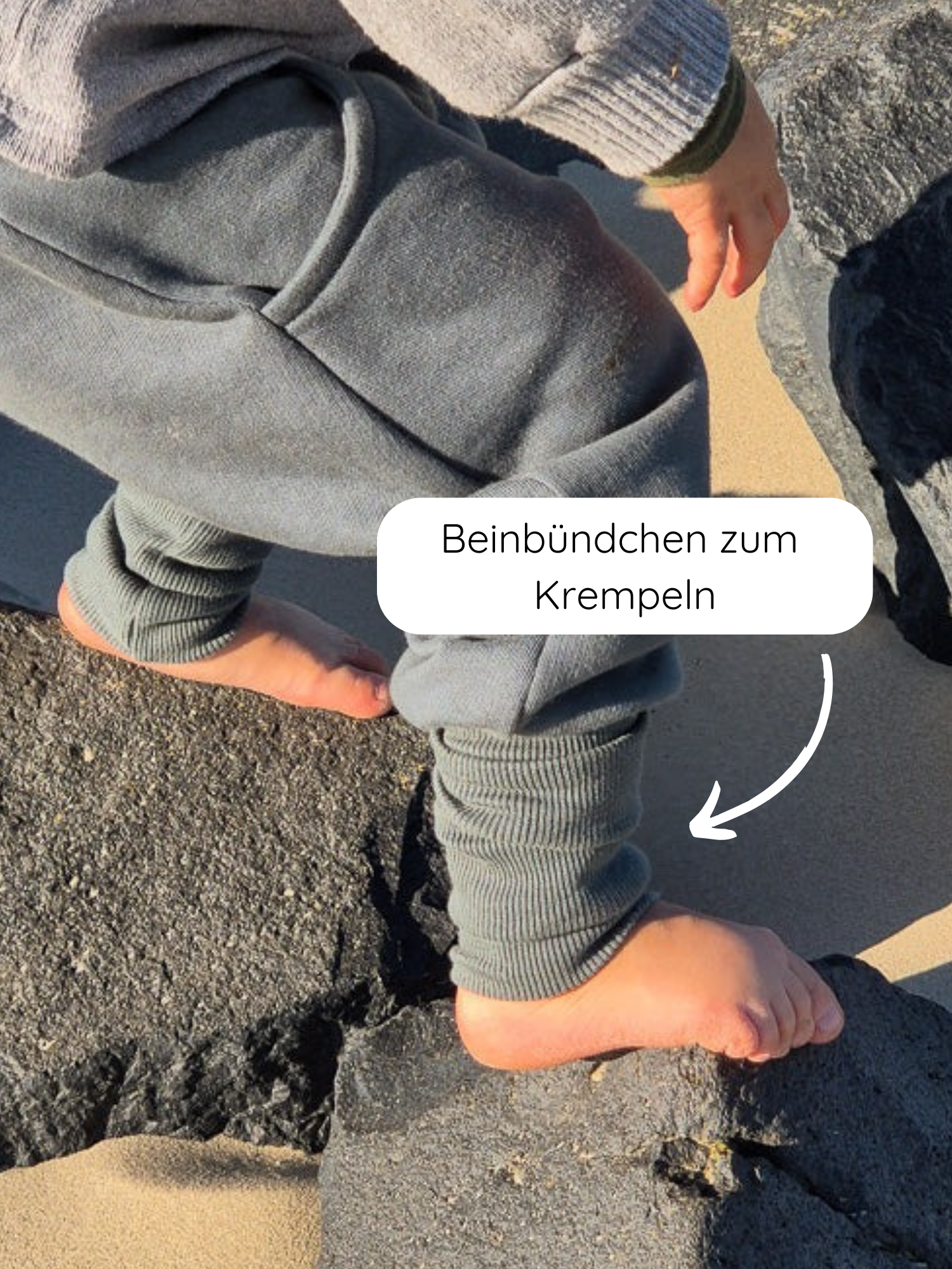 Ein Kind steht barfuß auf Vulkanstein und Sand, trägt die Knickerbocker Bio-Sweat - Taube mit hochgekrempelten, gerippten Beinabschlüssen, ideal für Spiel und Entdeckungstouren.