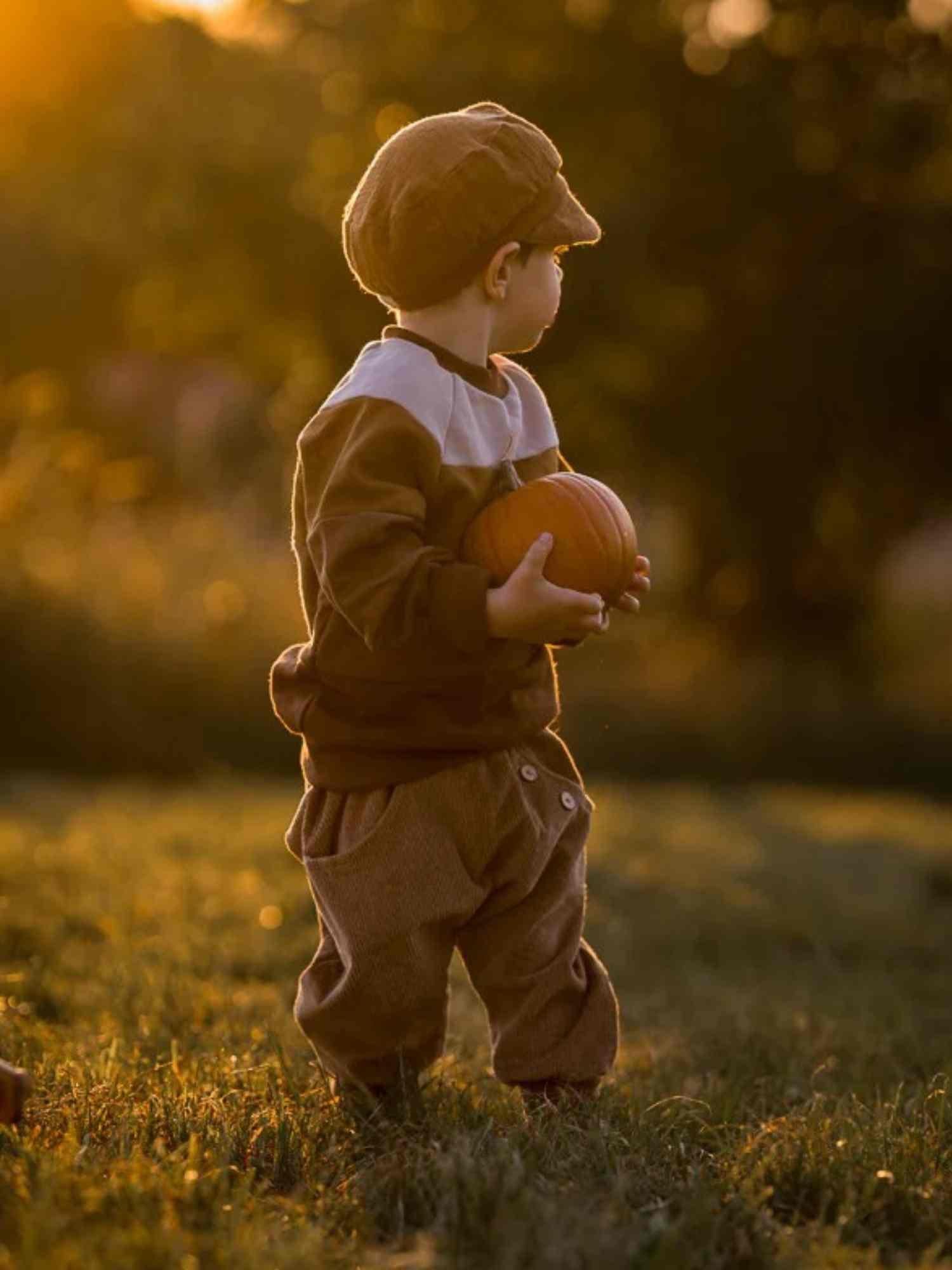 Ein Kleinkind steht in einem Feld bei Sonnenuntergang und hält einen Kürbis, gekleidet in Knickerbocker Breitcord Stretch - Weizen, ideal für kühle Tage und aktive Entdecker.