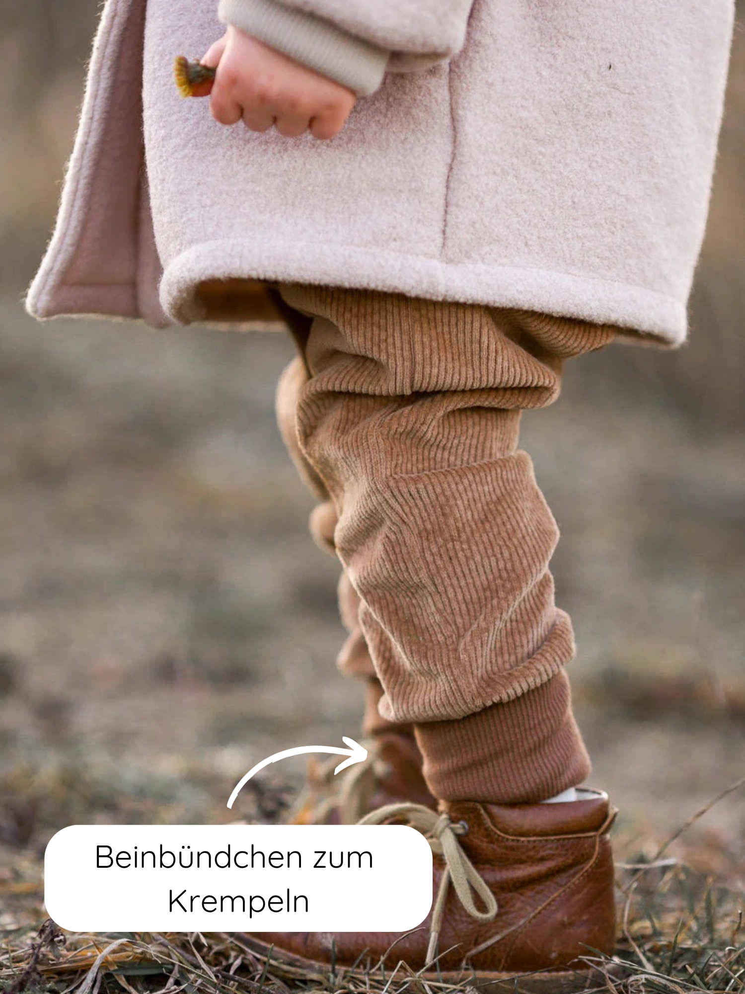 Knickerbocker Breitcord Stretch - Weizen: Nahaufnahme der Beine eines Kindes in hellbeigem Mantel, braunen Cordhosen mit aufgekrempeltem Beinbündchen und braunen Stiefeln, Hand hält kleinen Gegenstand.