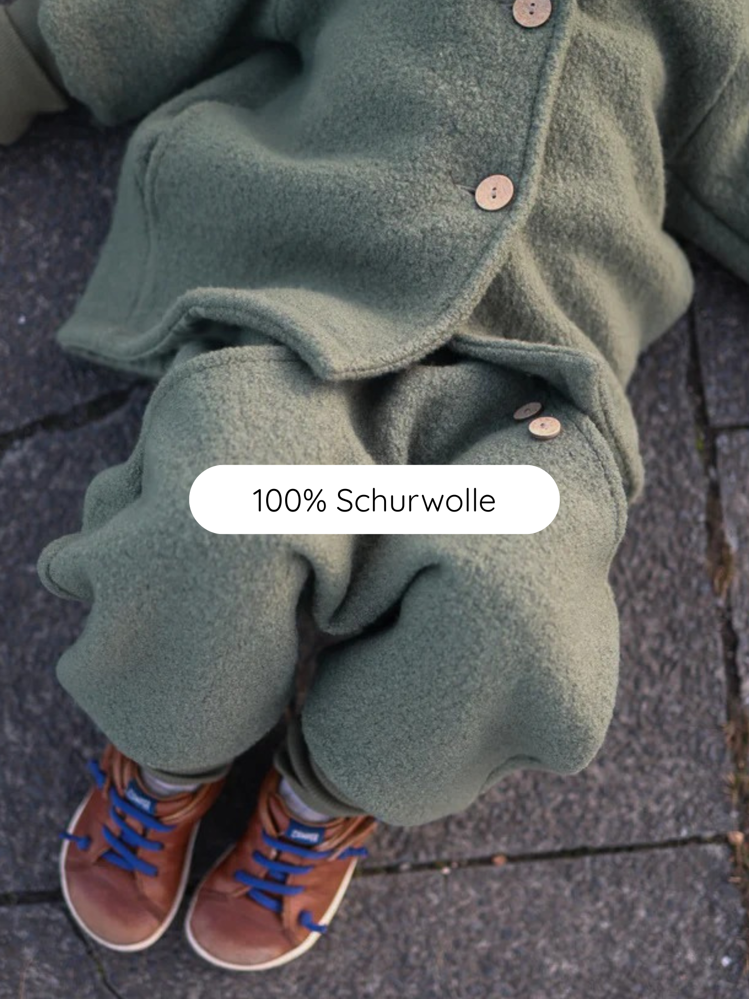 Knickerbocker Wollwalk - Pistazie: Ein Kind sitzt auf Pflaster, trägt ein mossgrünes Wolloutfit mit Holzknöpfen und braunen Sneakers mit blauen Schnürsenkeln. Ideal für kühles, nasses Wetter.