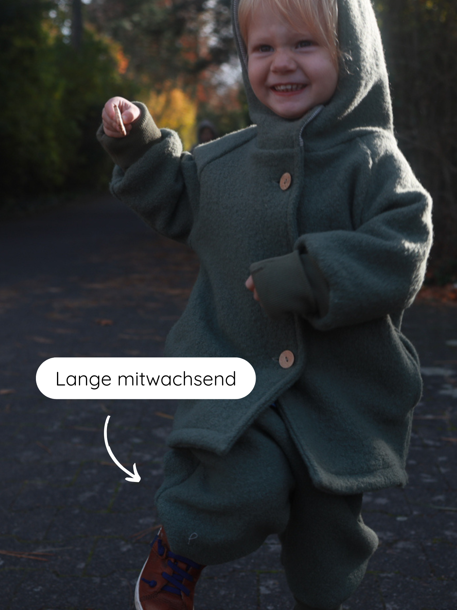 Kleinkind im Freien trägt Knickerbocker Wollwalk - Pistazie mit Kapuzenmantel, hält einen Stock, lächelt. Pfeil zeigt auf Hosenbein, betont lange mitwachsend, ideal für Herbst bis Frühling.