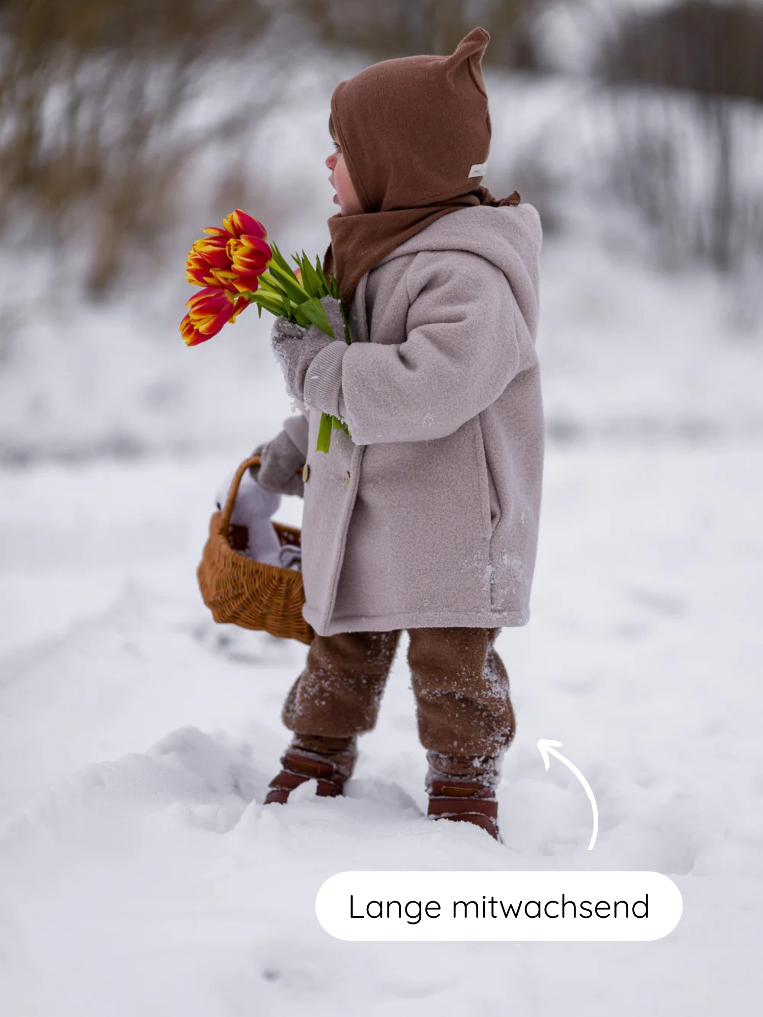 Ein Kind steht im Schnee, trägt die Knickerbocker Wollwalk - Schoko mit Kängurutaschen und Bio-Baumwollbündchen, ergänzt durch braune Stiefel. Ein Pfeil zeigt auf die längenverstellbaren Hosenbeine.
