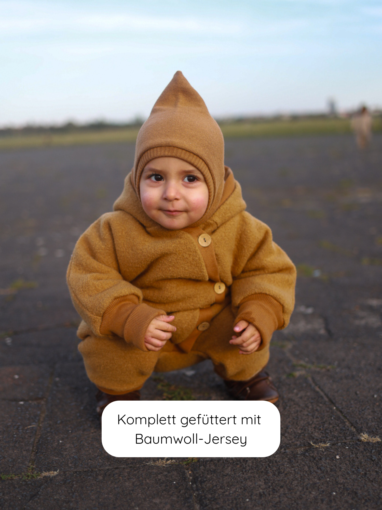 Kleinkind im Kuschelanzug Wollwalk - Honig mit spitzer Kapuze und Holzknöpfen, aus 100% Schurwolle, komplett gefüttert mit Baumwoll-Jersey, auf gepflastertem Boden, passende braune Stiefel tragend.