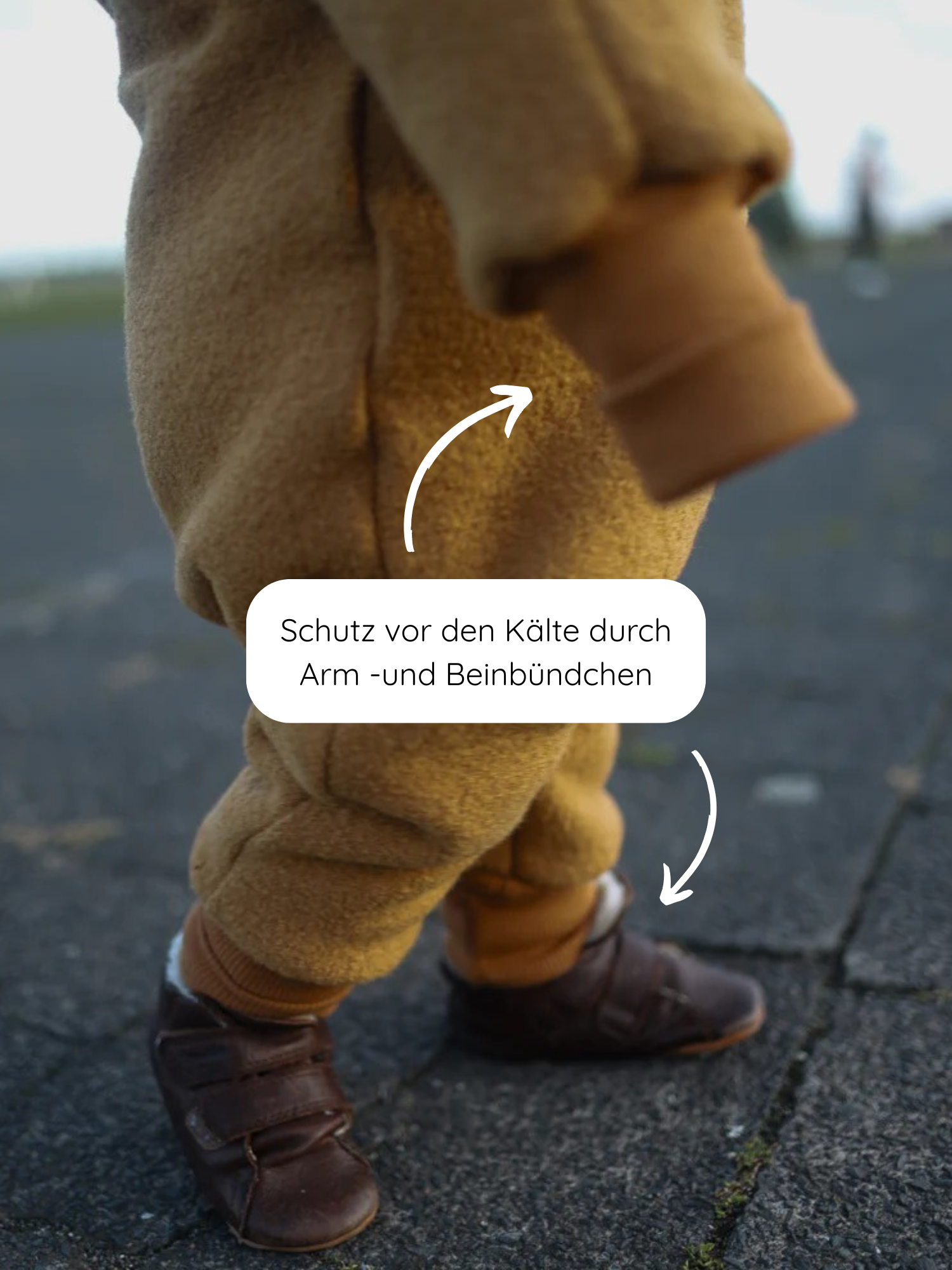 Kleinkind in Kuschelanzug Wollwalk - Honig mit elastischen Arm- und Beinbündchen, die vor Kälte schützen. Dazu dunkle Lederschuhe mit Klettverschluss auf Pflastersteinboden.