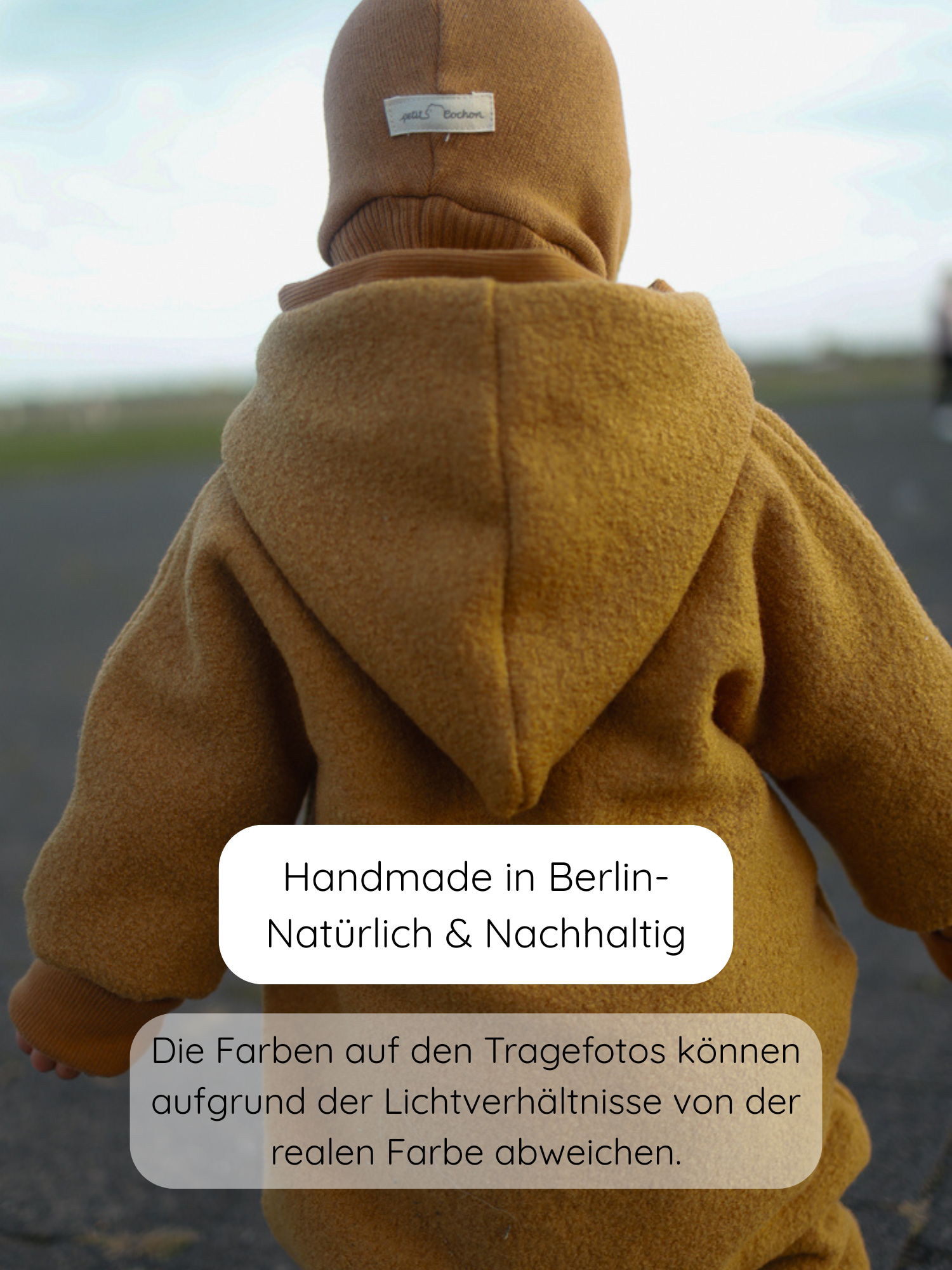 Kleinkind von hinten in einem Kuschelanzug Wollwalk - Honig auf einem Weg. Der Anzug ist einteiliger, warmer Overall mit Kapuze und Balaclava, handgefertigt in Berlin, ideal für kühle Jahreszeiten.