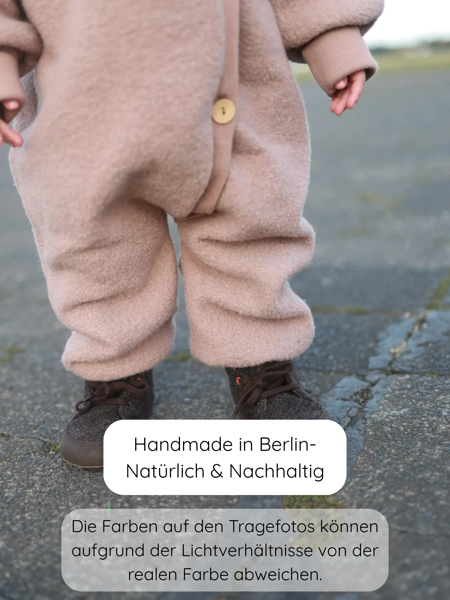 Kleinkind trägt Kuschelanzug Wollwalk - Puder, einen flauschigen, einteiligen Overall aus 100% Schurwolle mit Baumwollfutter, kombiniert mit dunkelbraunen Schnürstiefeln, auf Pflasterstein stehend.