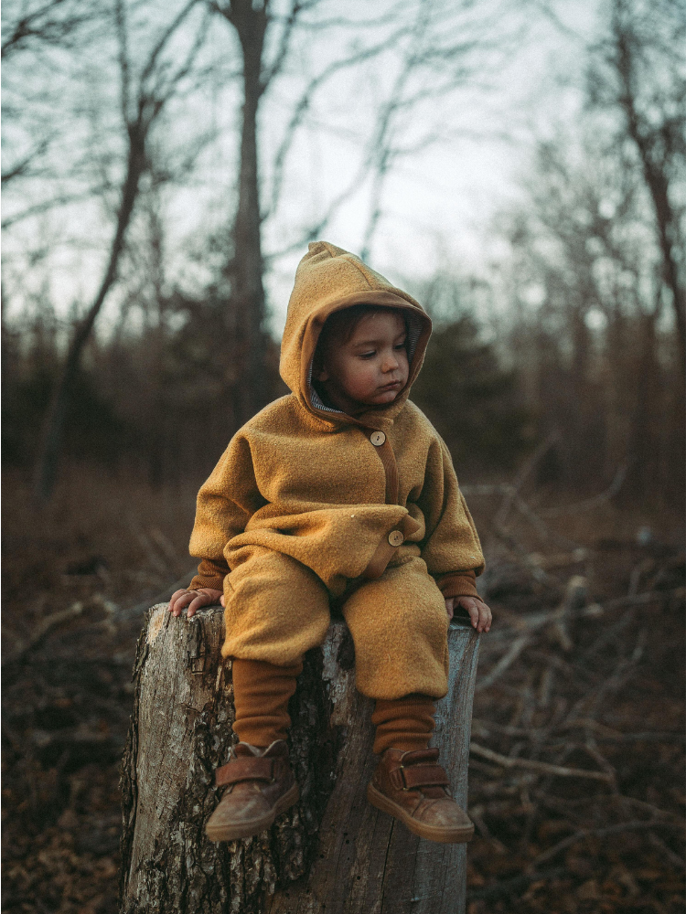 Kleinkind sitzt auf Baumstumpf, trägt den Kuschelanzug Wollwalk - Honig aus 100% Schurwolle mit Bio-Jersey-Futter. Der Anzug bietet Bewegungsfreiheit und Wärme, ideal für kühle Spaziergänge.