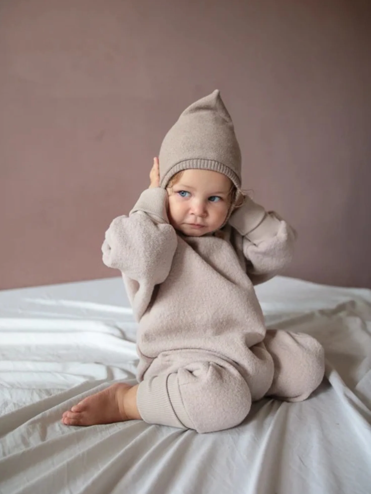 Ein Kleinkind sitzt auf einem Bett und trägt den Kuschelanzug Wollwalk - Sand, ein beiges Strickoutfit mit Mütze, ideal für kühle Tage, aus 100% Schurwolle mit Baumwollfutter.