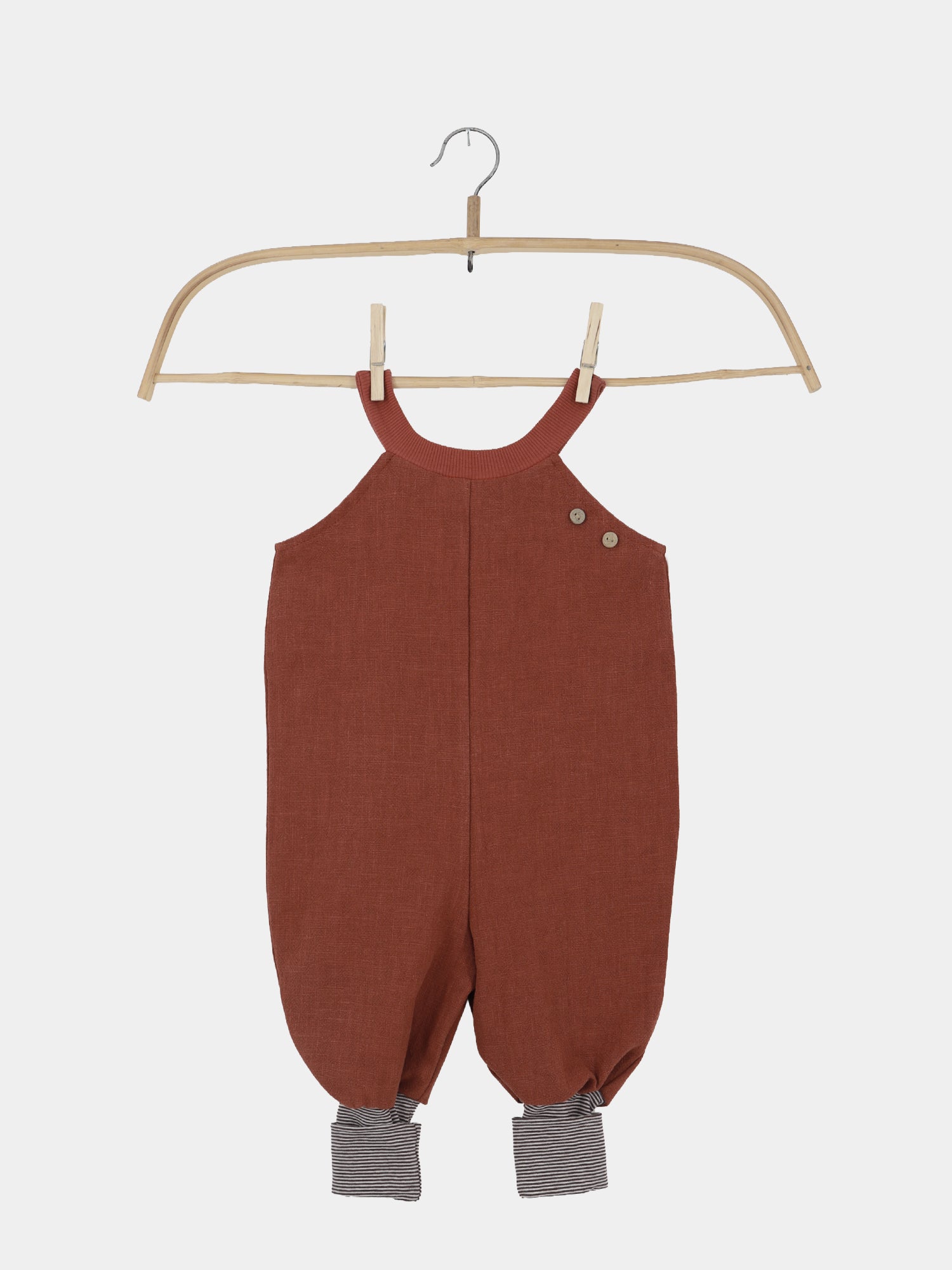 Schlupflatzhose Leinen - Ziegel: Rustfarbene, ärmellose Baby-Latzhose mit Knöpfen auf einer Schulter, tieferem Schritt und gestreiften Bündchen an den Beinen, auf einem Holzkleiderbügel, vor weißem Hintergrund.