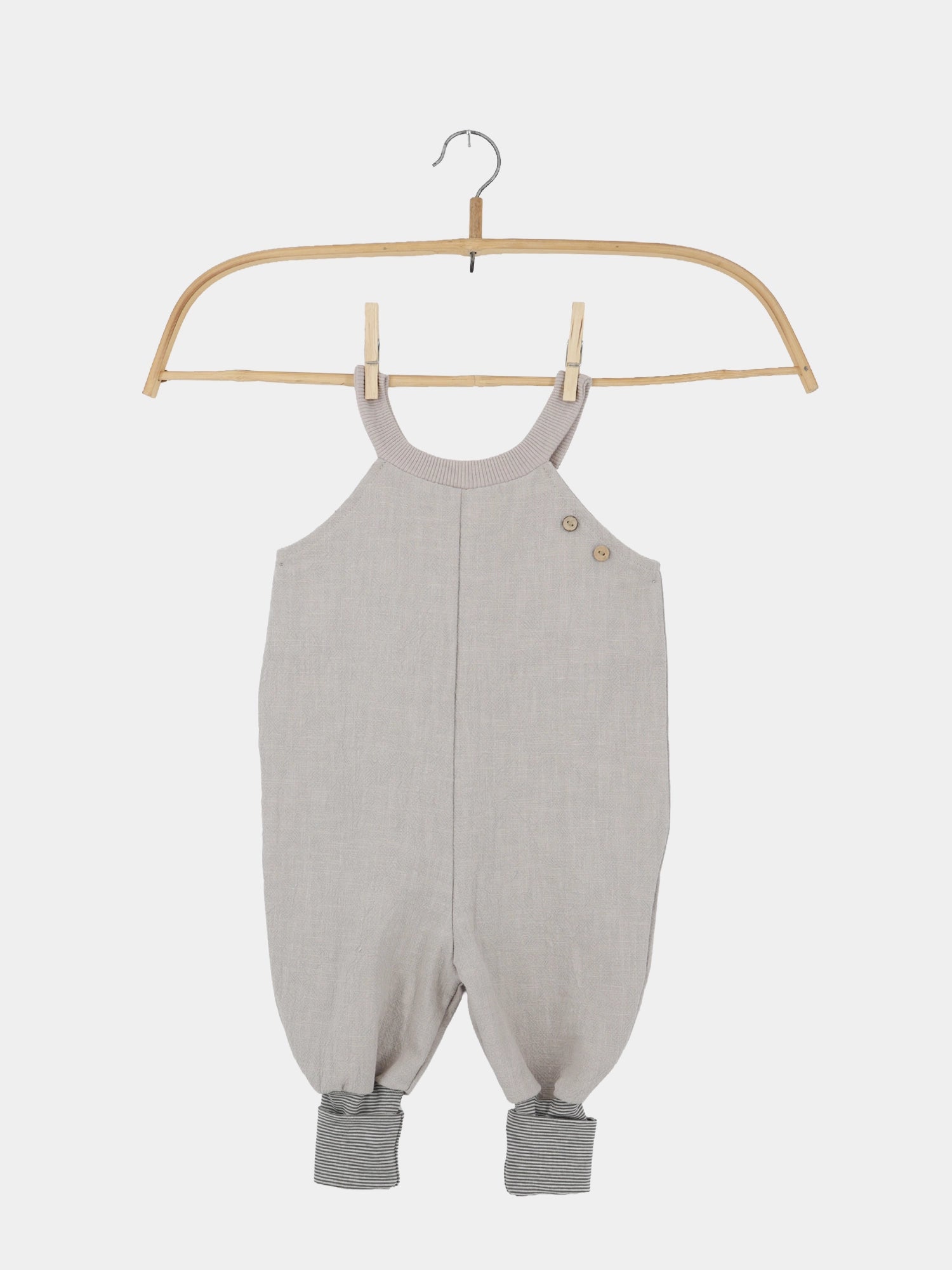 Schlupflatzhose Leinen - Sand: Ein hellgrauer, ärmelloser Baby-Strampler mit zwei Holzknöpfen, geripptem Kragen und gestreiften Knöchelbündchen, hängend an einem Holzkleiderbügel mit Wäscheklammern.