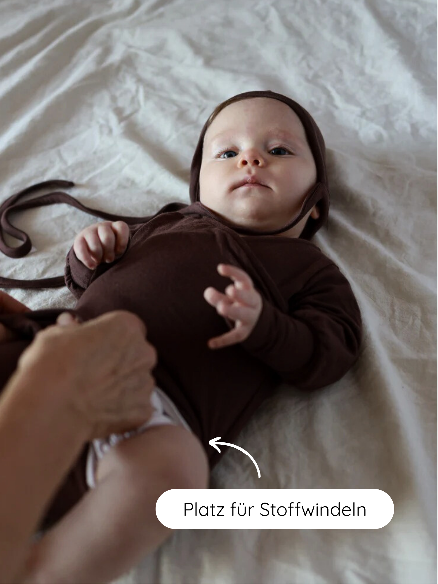 Ein Baby trägt den Loopbody Bambusviskose - Zartbitter mit passender Haube, während eine erwachsene Hand eine Stoffwindel in den großzügigen, windelgerechten Schnitt des Bodys einpasst.