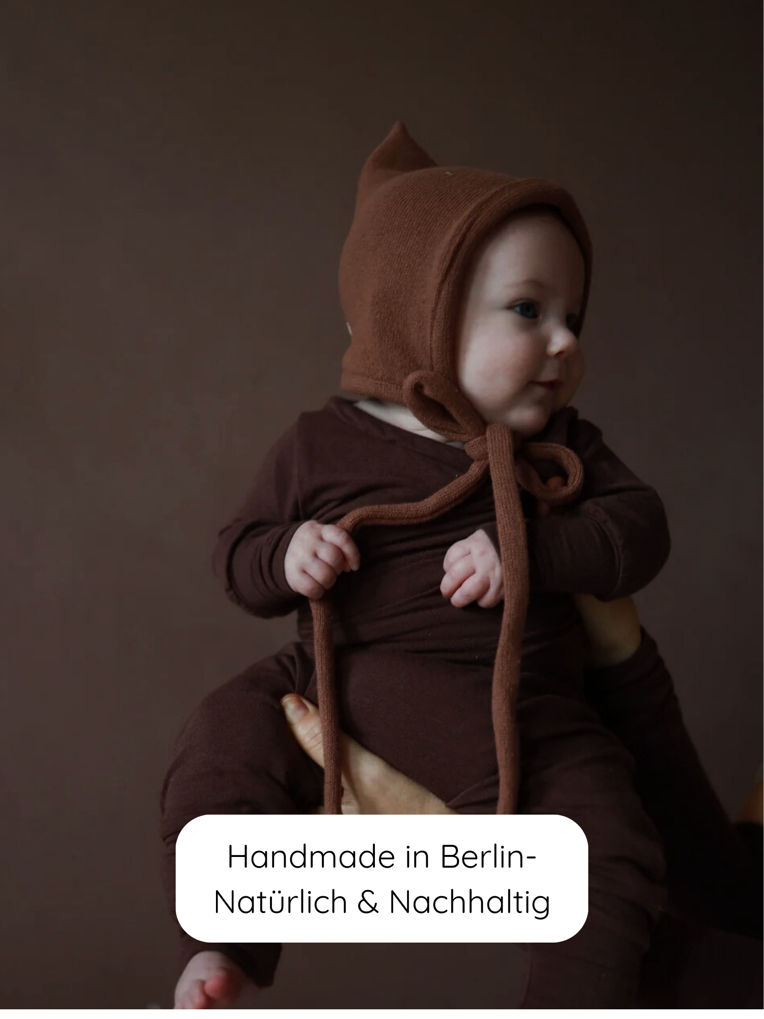 Ein Baby trägt den Loopbody Bambusviskose - Zartbitter, bestehend aus einem braunen Strickset mit Langarmshirt, passender Hose und Bonnet, gehalten vor einem braunen Hintergrund, mit dem Label „Handmade in Berlin“.