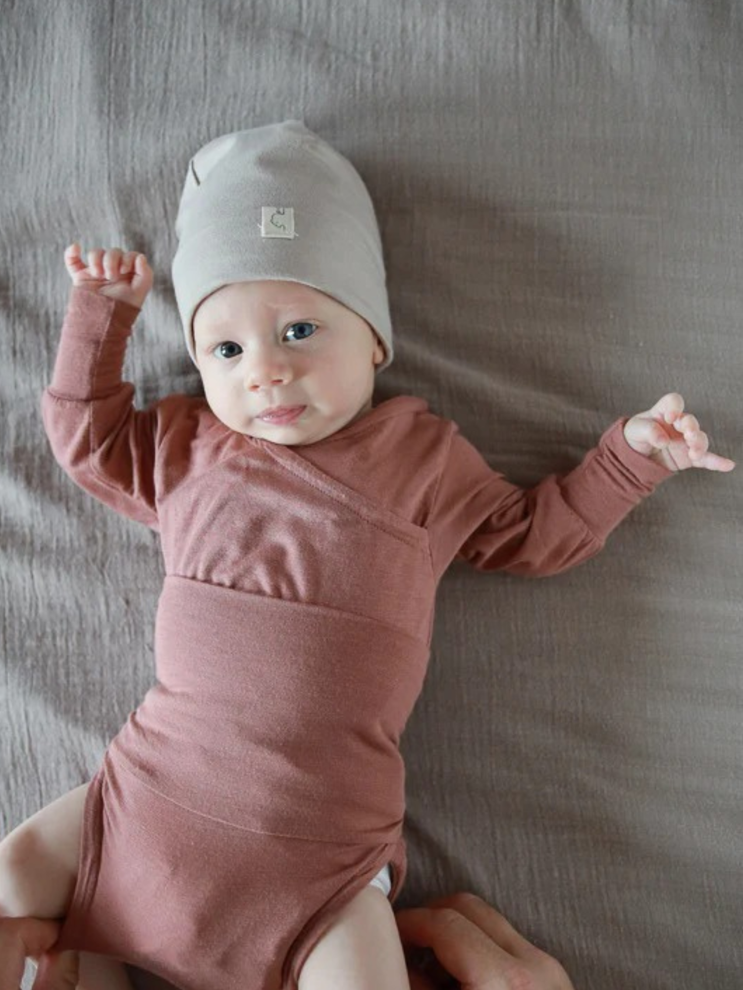 Baby in einem Loopbody Kaschmir-Mix - mitwachsender Babybody - Zimt, liegt auf einer weichen Unterlage. Es trägt einen grauen Strickhut, ein Arm ist angewinkelt, der andere ausgestreckt.