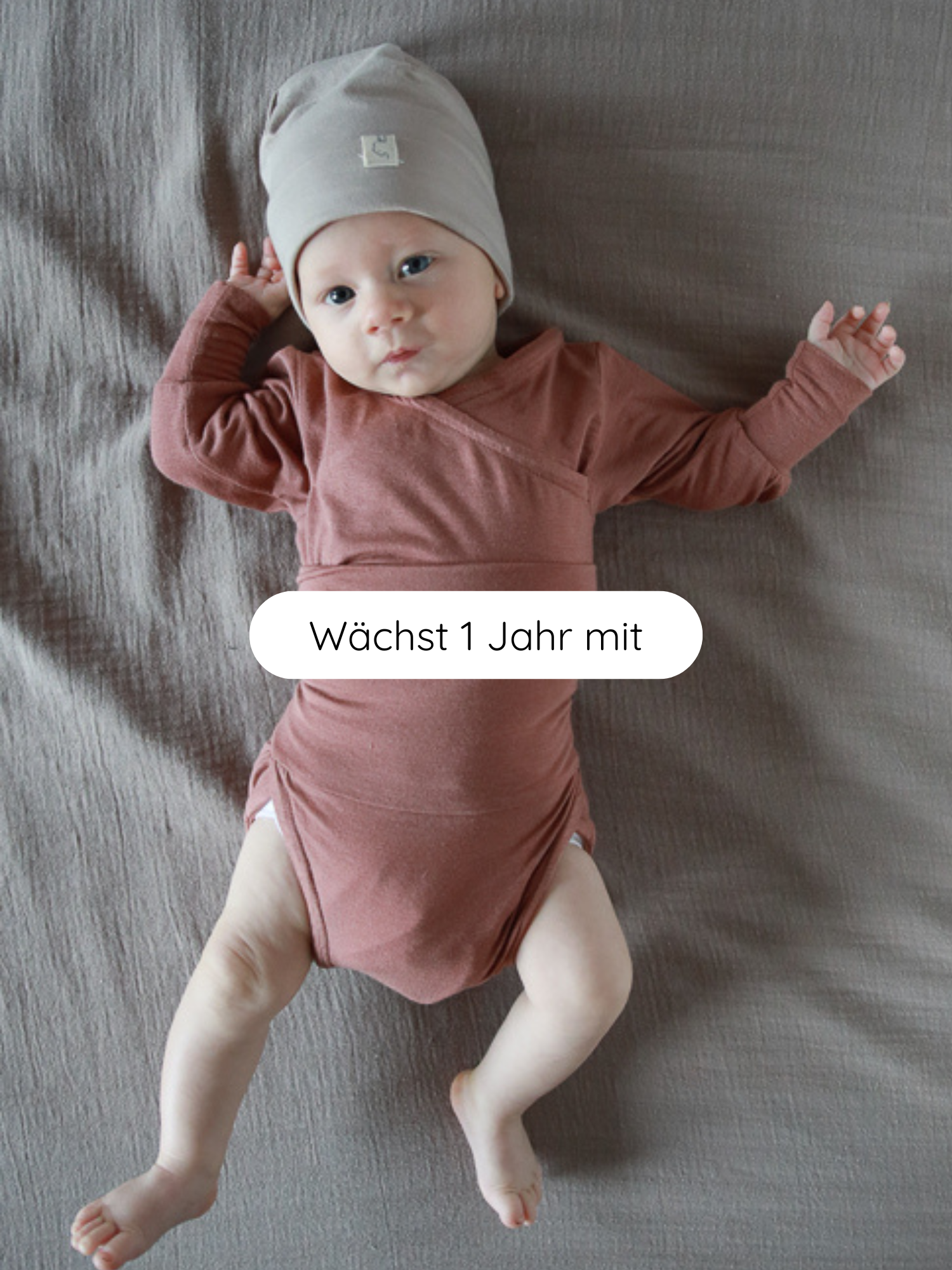Baby in einem Loopbody Kaschmir-Mix - mitwachsender Babybody - Zimt, mit Wickeldesign und grauer Mütze auf weicher Unterlage liegend. Text „Wächst 1 Jahr mit“ über dem Bild.