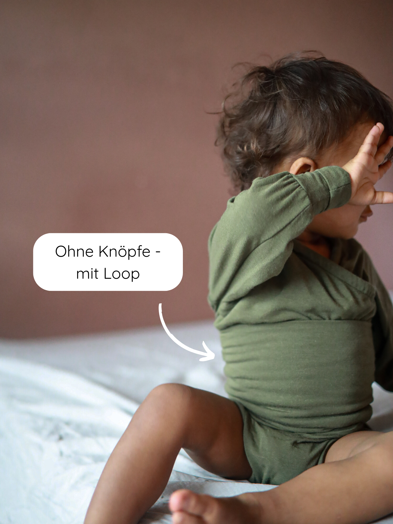 Kleinkind sitzt auf Bett im Loopbody Kaschmir-Mix - Avocado, ohne Knöpfe, mit Loop-Verschluss am Bauch, hält eine Hand zum Gesicht.