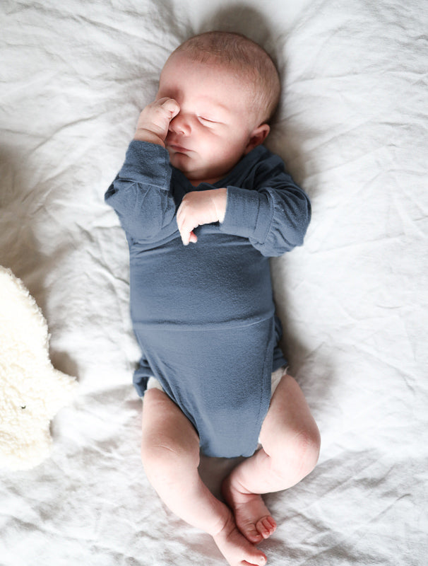 Ein Neugeborenes liegt auf einem weißen Bettlaken und trägt den Loopbody Kaschmir-Mix - Blaubeere, einen langärmeligen, weichen Body ohne Knöpfe, ideal für empfindliche Babyhaut.