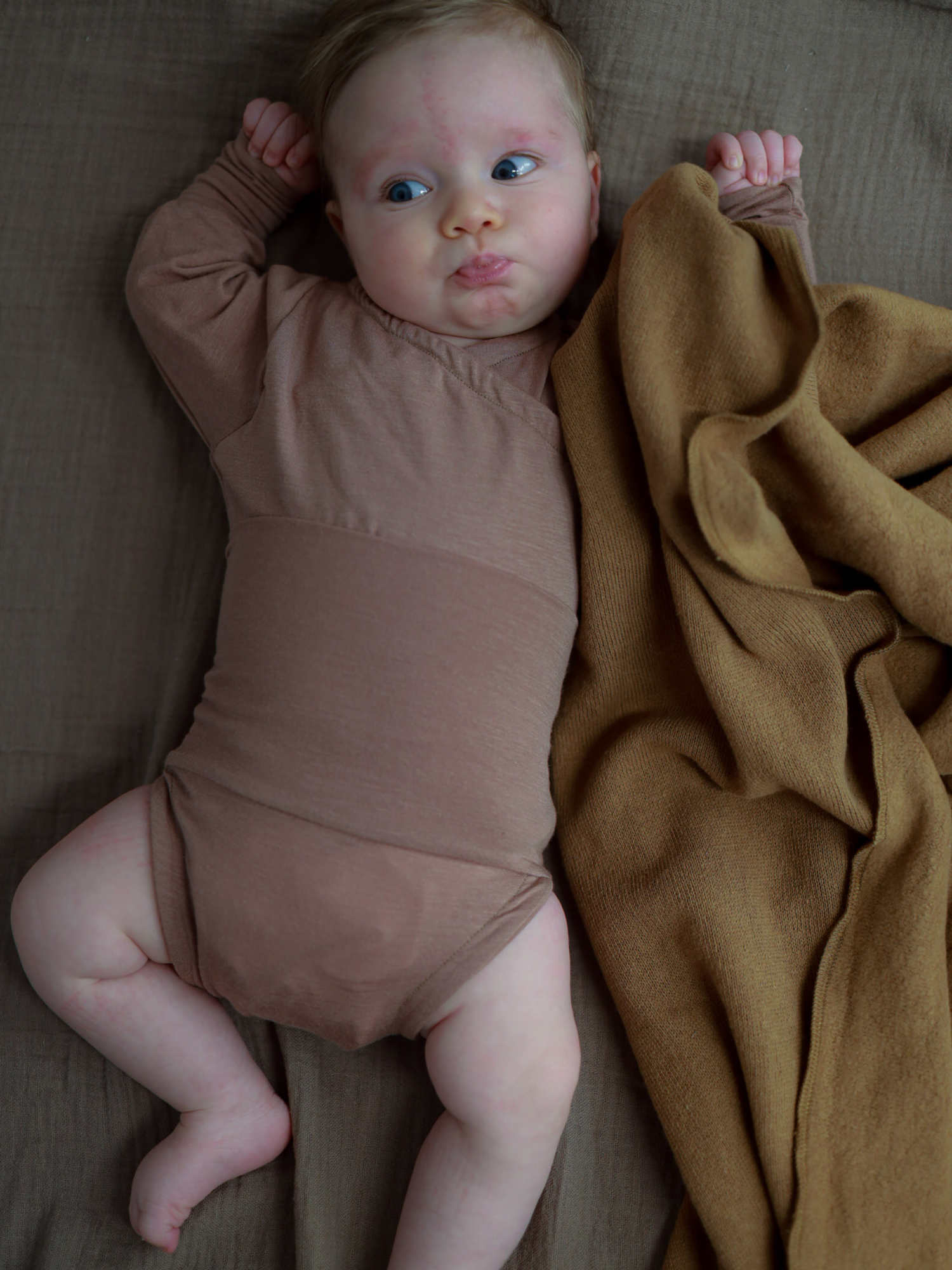 Baby in einem Langarm-Loopbody Kaschmir-Mix - Puder, teilweise von einer weichen Decke bedeckt, schaut zur Seite. Der Body umschließt sanft den Bauch und bietet Komfort und Wärme.