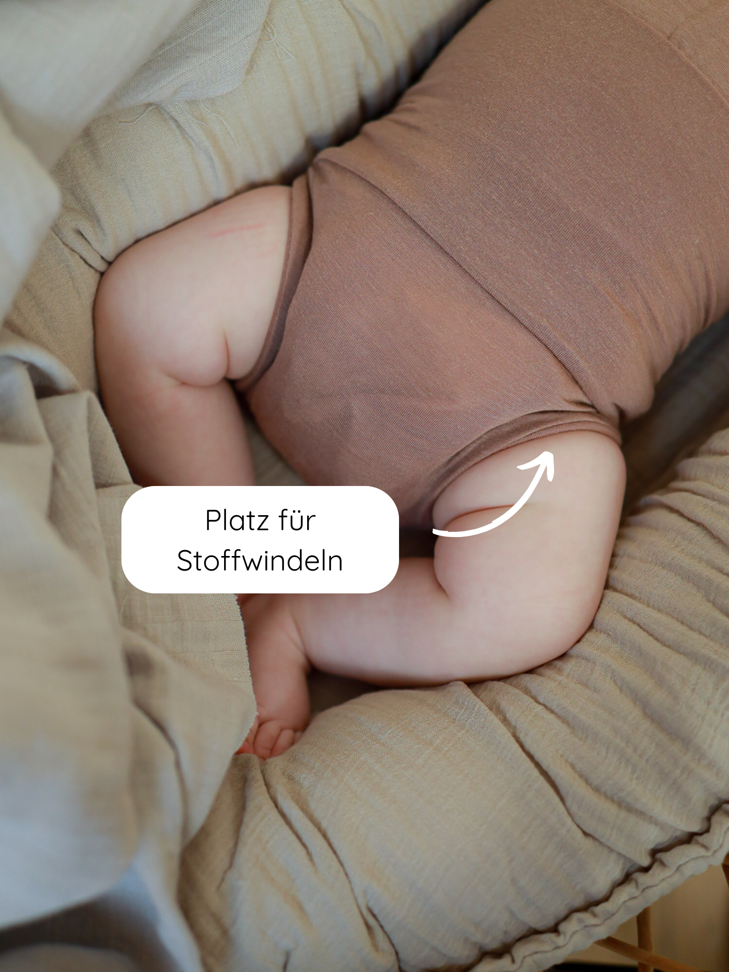 Ein Baby in einem Loopbody Kaschmir-Mix - Puder liegt im beigen Babynest, mit einem Pfeil auf den Po für Stoffwindeln. Der Body ist supersoft, atmungsaktiv und mitwachsend.