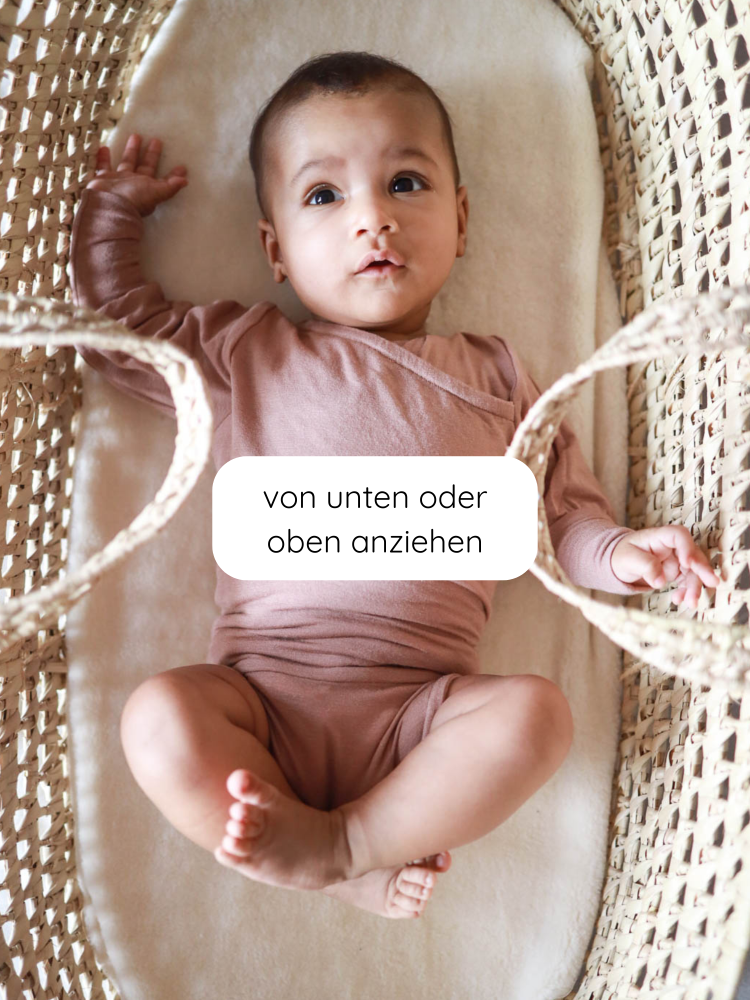 Ein Säugling liegt in einem gewebten Moses-Korb auf einer weichen Matratze, gekleidet in einen Loopbody Kaschmir-Mix - Puder, der durch ein innovatives, knopf- und nahtloses Design besticht.