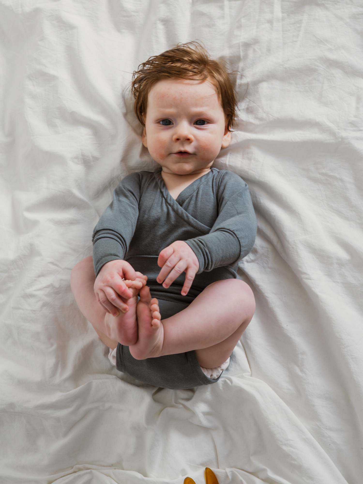 Baby in grauem Loopbody Kaschmir-Mix - Taube liegt auf weißem Bett, hält seine Füße. Der Body umhüllt sanft den Bauch, bietet Komfort und wächst mit. Gelber Schnuller am Rand sichtbar.