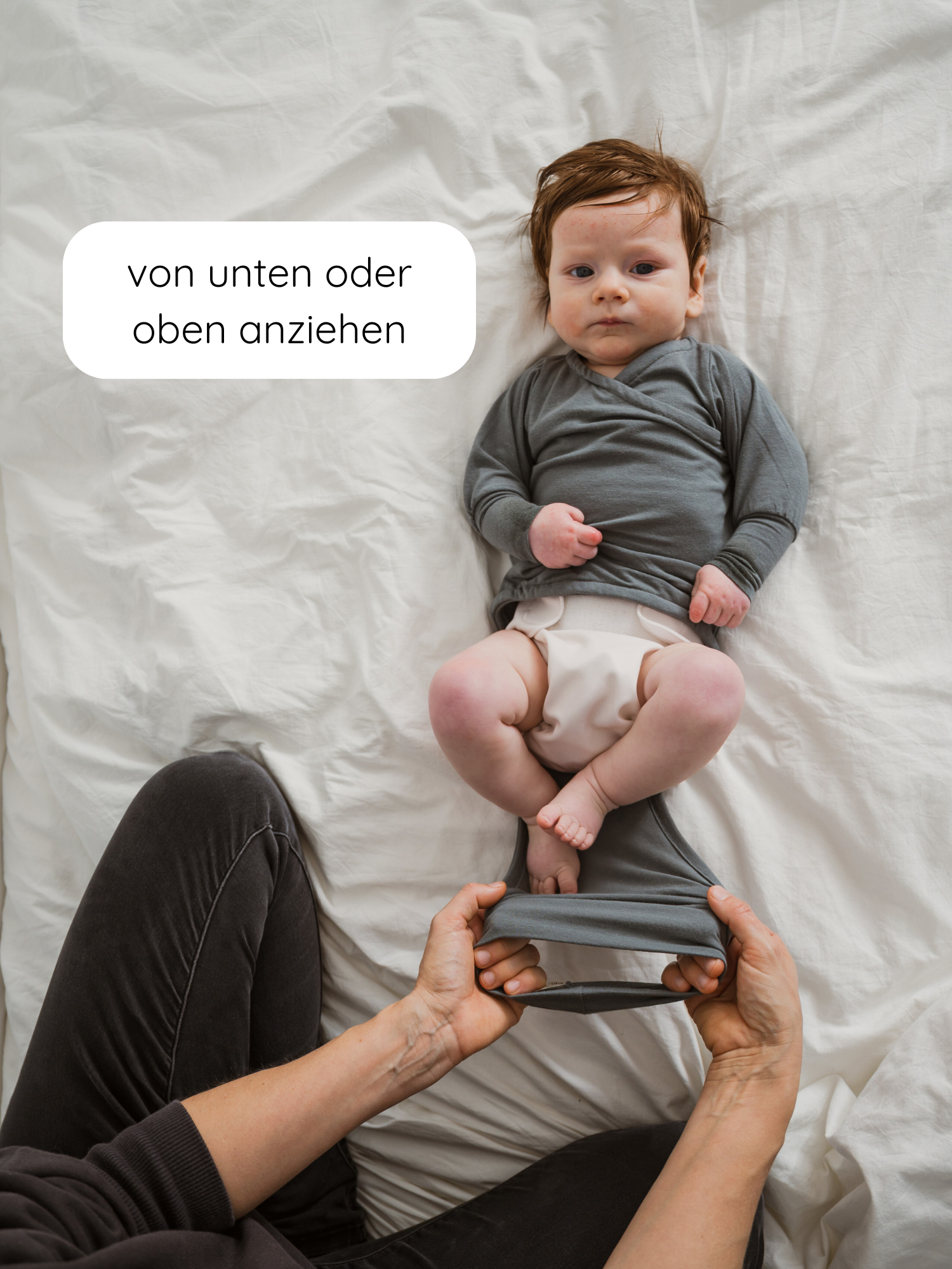 Loopbody Kaschmir-Mix - Taube: Ein Baby liegt in einem grauen Wickeloberteil und Windel, während Hände einen onesie artiges Kleidungsstück über die Beine ziehen, unterstützt durch den einzigartigen Loop-Design für einfaches Anziehen.