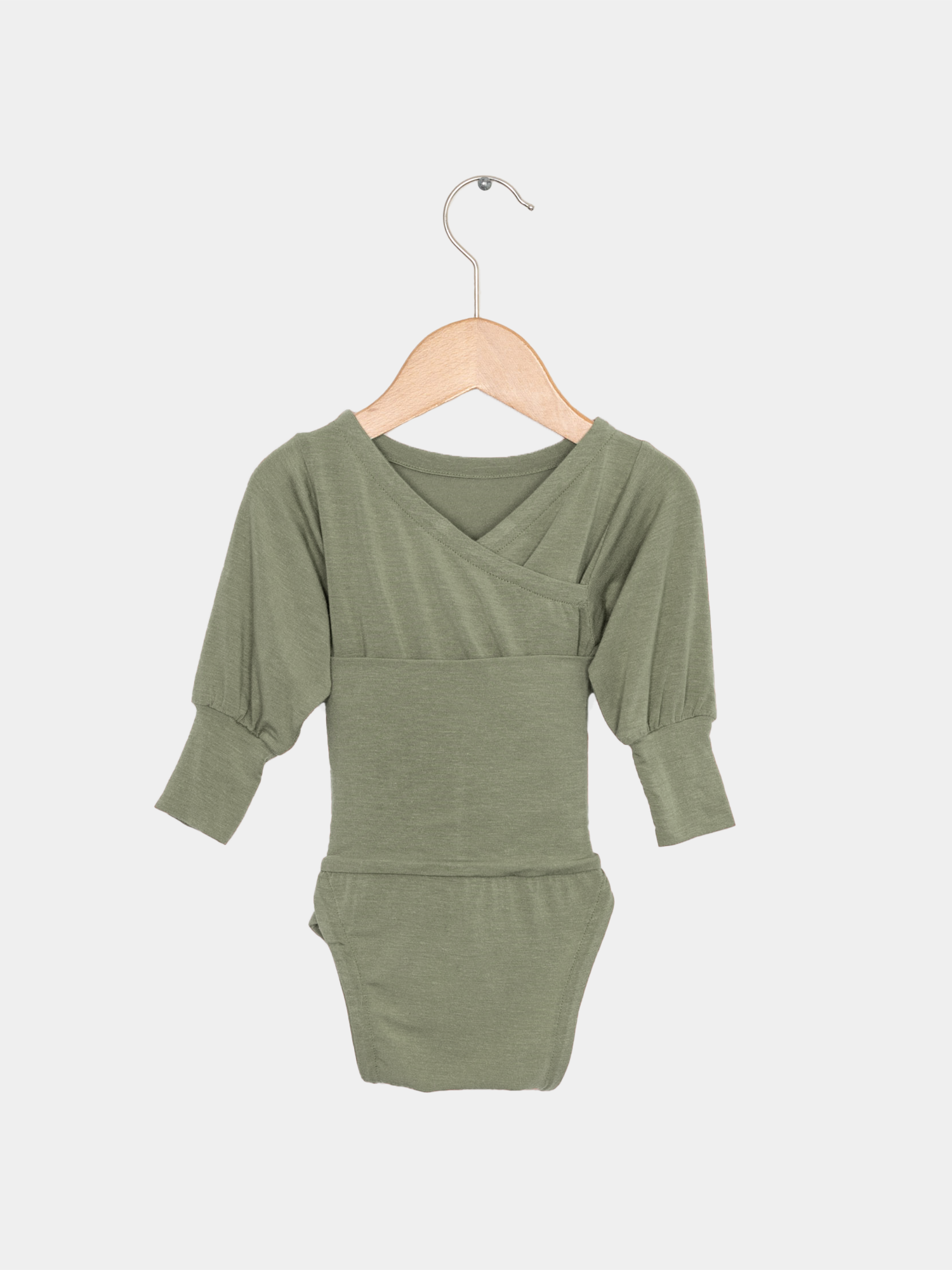 Loopbody Kaschmir-Mix - Pistazie: Ein olivgrüner Langarm-Babybody mit einzigartigem Crossover-Design und Loop-Detail, das extra Wärme bietet. Hängend an einem einfachen Holzbügel.