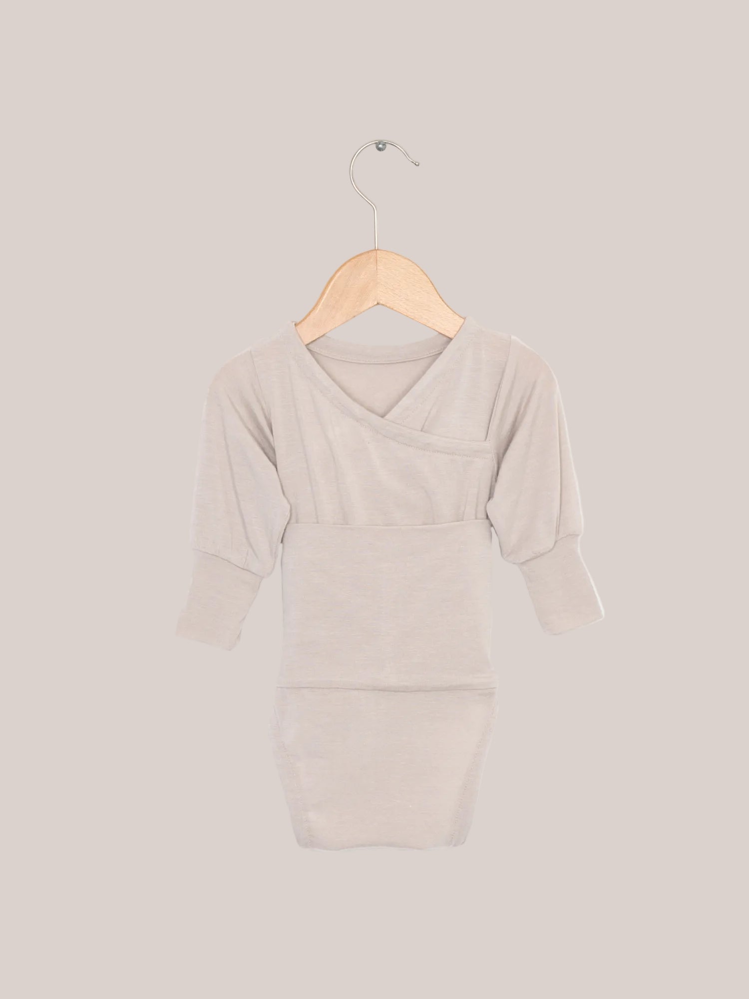 Body para bebé de mezcla de cachemir Loopbody - Arena: un body para bebé sin botones en una percha de madera. El suave body de manga larga tiene un lazo especial para ponérselo fácilmente y ofrece un calor adicional. Canastilla para bebé y regalo de nacimiento.