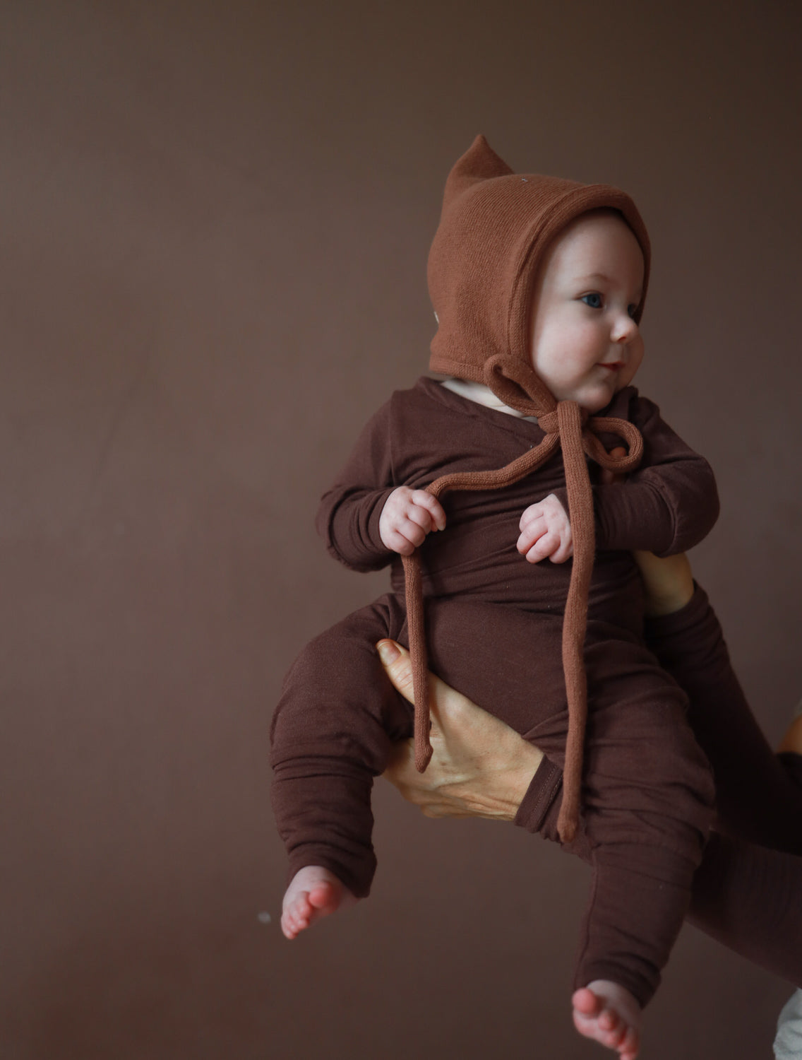Ein Baby, gehalten von Erwachsenen, trägt den Loopbody Bambusviskose - Zartbitter in Schokoladenbraun, inklusive langer Ärmel und spitzer Mütze, mit neugierigem Blick zur Seite.