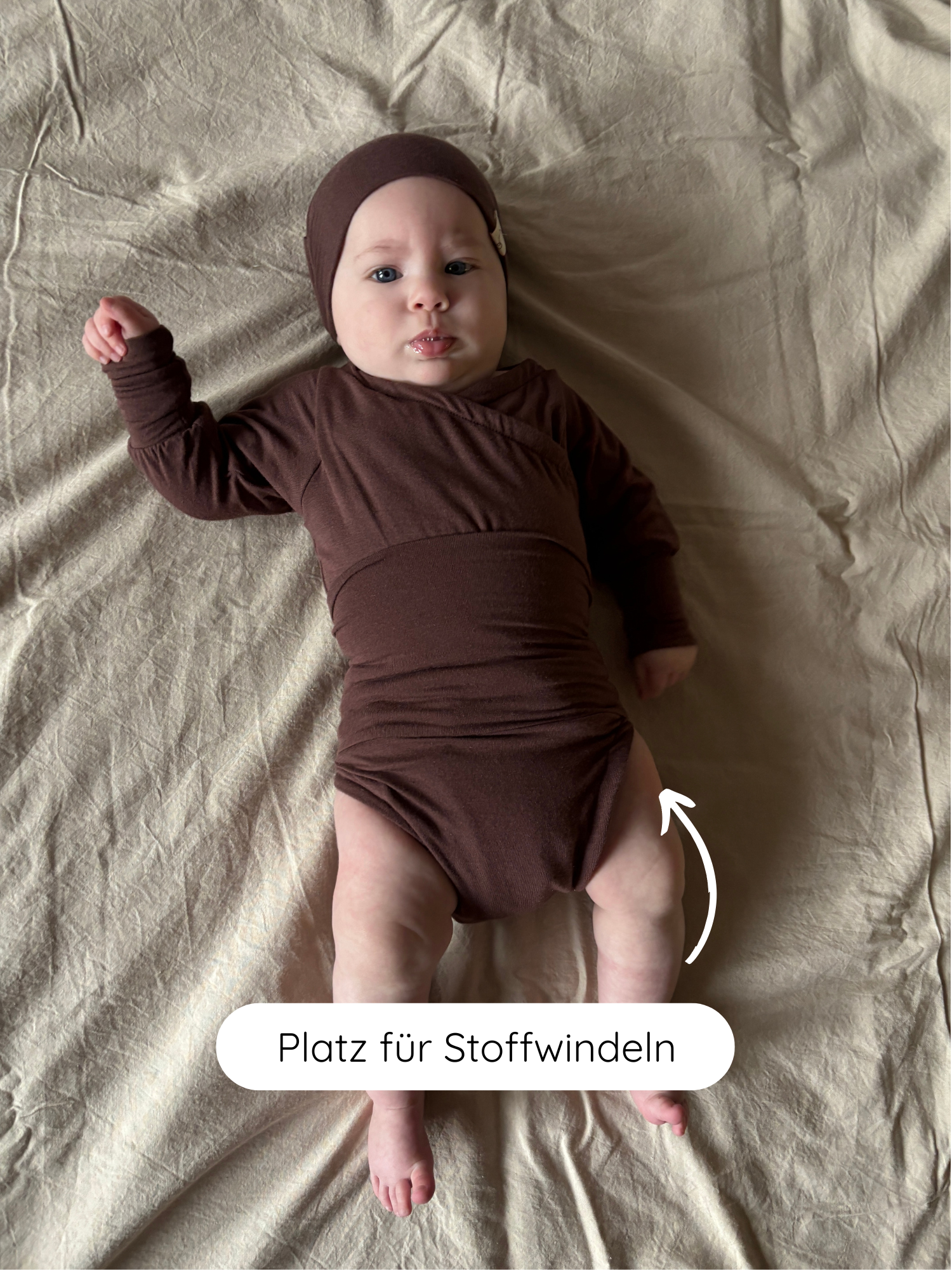Baby in einem Loopbody Kaschmir-Mix - Malve und passender Haube. Der Body ist weich, mitwachsend und bietet Platz für Stoffwindeln. Ein weißer Pfeil zeigt auf den Hüftbereich.