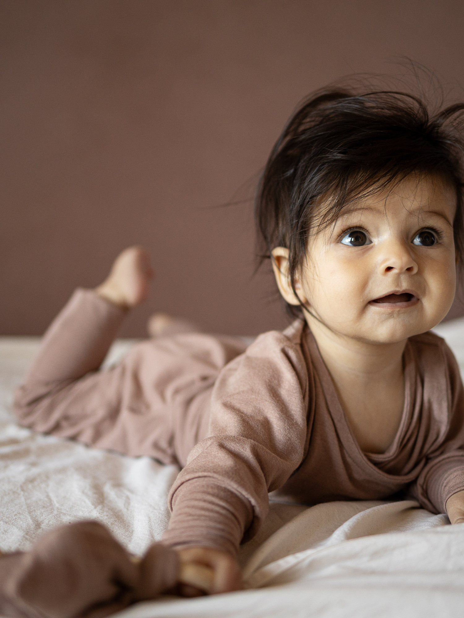 Ein Baby liegt auf dem Bauch auf einer weichen Oberfläche und trägt den Loopstrampler Kaschmir-Mix - Puder, ohne Knöpfe, mit langen Ärmeln und umklappbaren Bündchen für wärmende Socken.