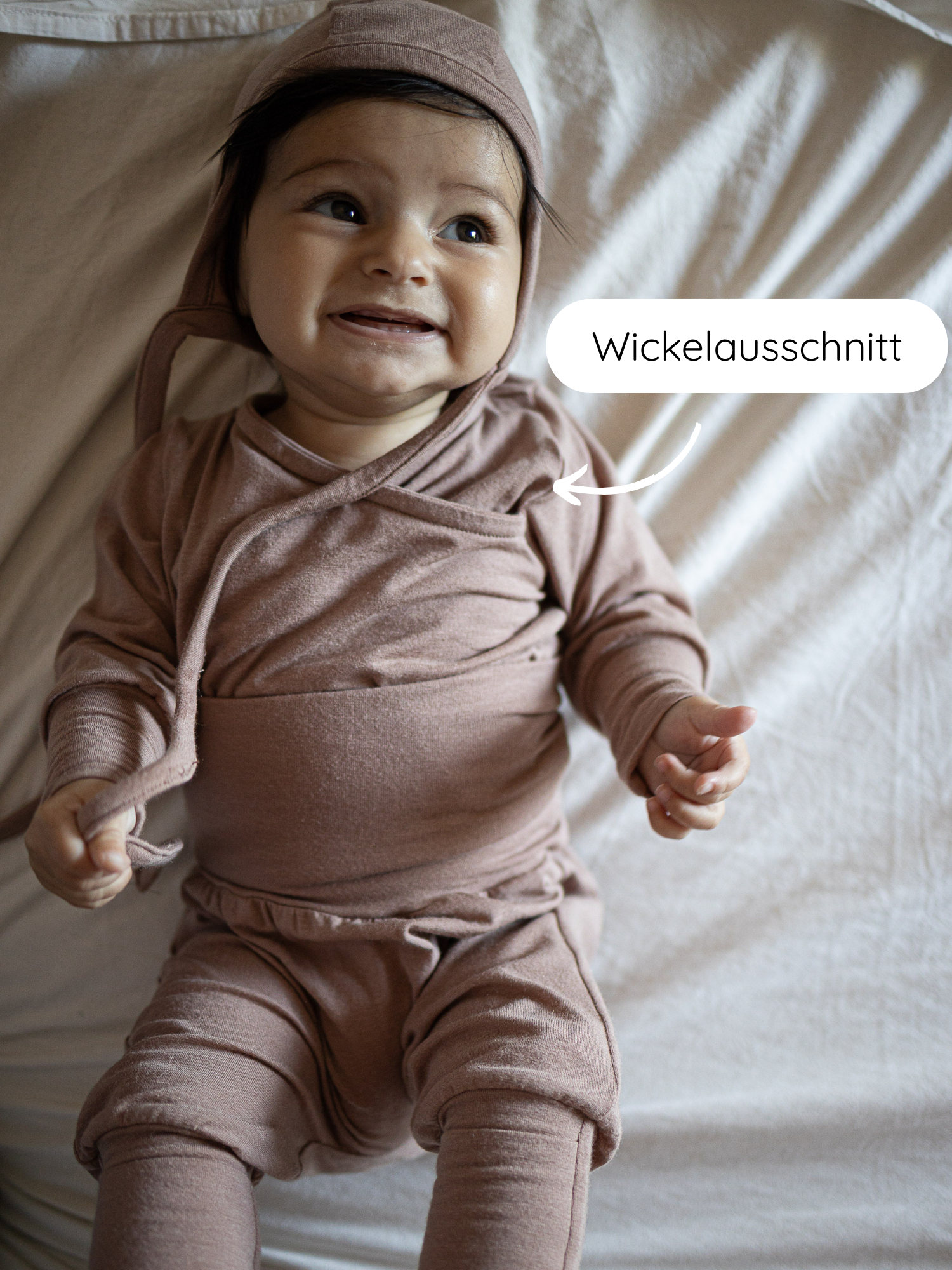 Ein lächelndes Baby trägt den Loopstrampler Kaschmir-Mix - Puder, bestehend aus einem weichen, zweiteiligen Outfit mit Wickelausschnitt und einer dazugehörigen Mütze, auf einer hellen Decke liegend.