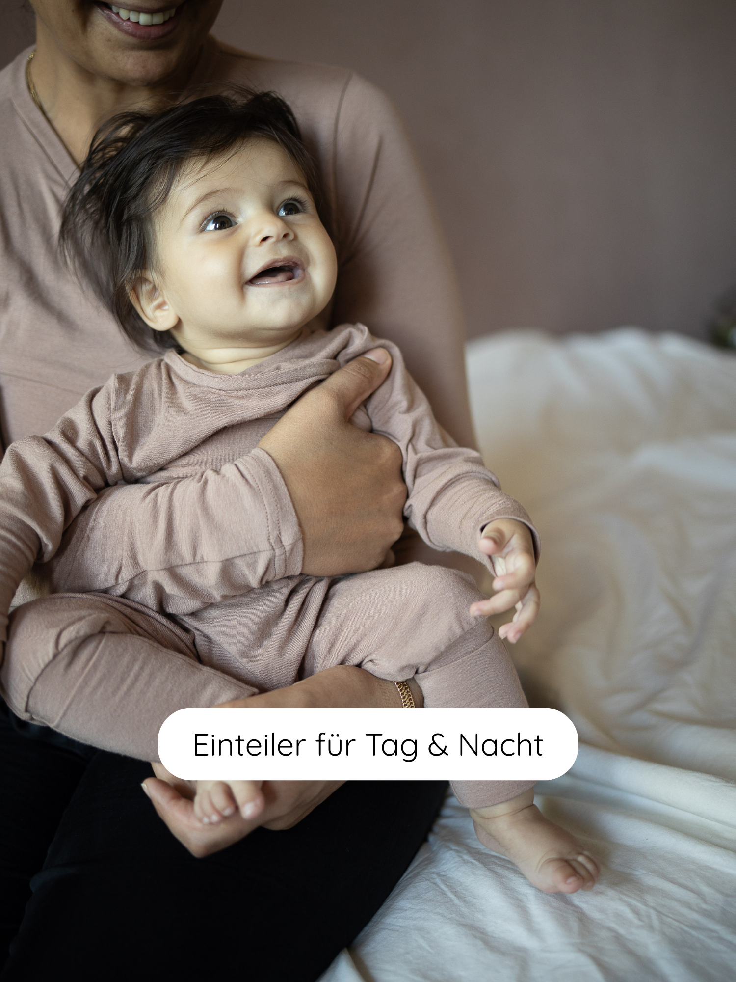 Ein lächelndes Baby im Loopstrampler Kaschmir-Mix - Puder wird in den Armen eines Erwachsenen gehalten, beide tragen beige Einteiler. Text: Einteiler für Tag & Nacht suggeriert einen vielseitigen Strampler.