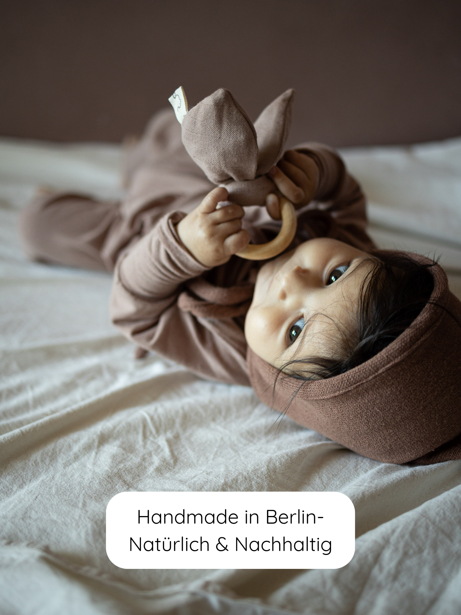 Kleines Baby in weichem, braunem Loopstrampler Kaschmir-Mix - Puder mit passender Mütze, hält einen Holz-Beißring. Unten Label: Handmade in Berlin – Natürlich & Nachhaltig.