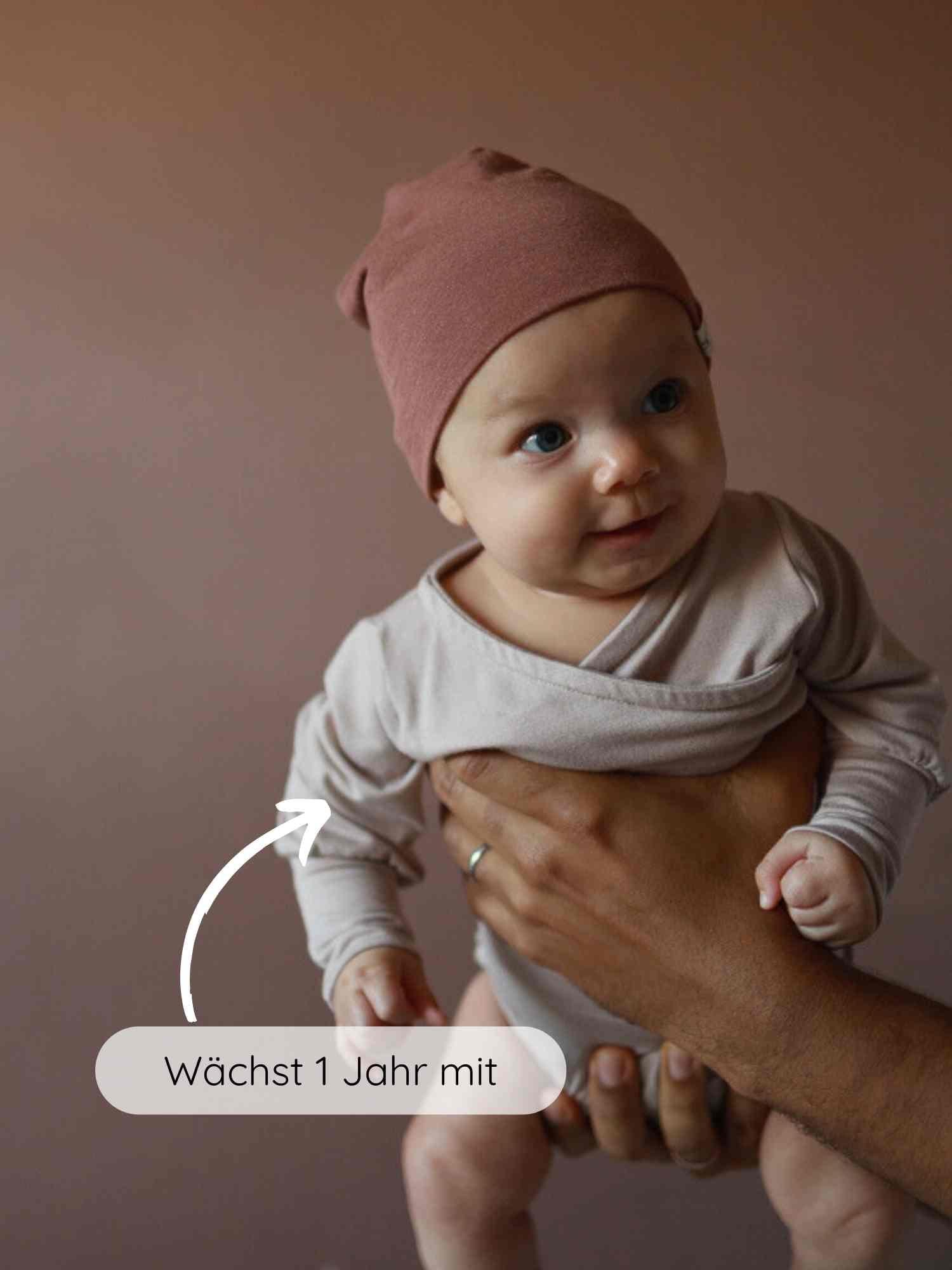 Loopbody Bambusviskose - Cremepuder: Ein Baby trägt einen beigen, langärmeligen Wickelbody und eine rostfarbene Mütze, gehalten von Erwachsenen. Ein Pfeil zeigt auf den Body mit dem Text Wächst 1 Jahr mit.