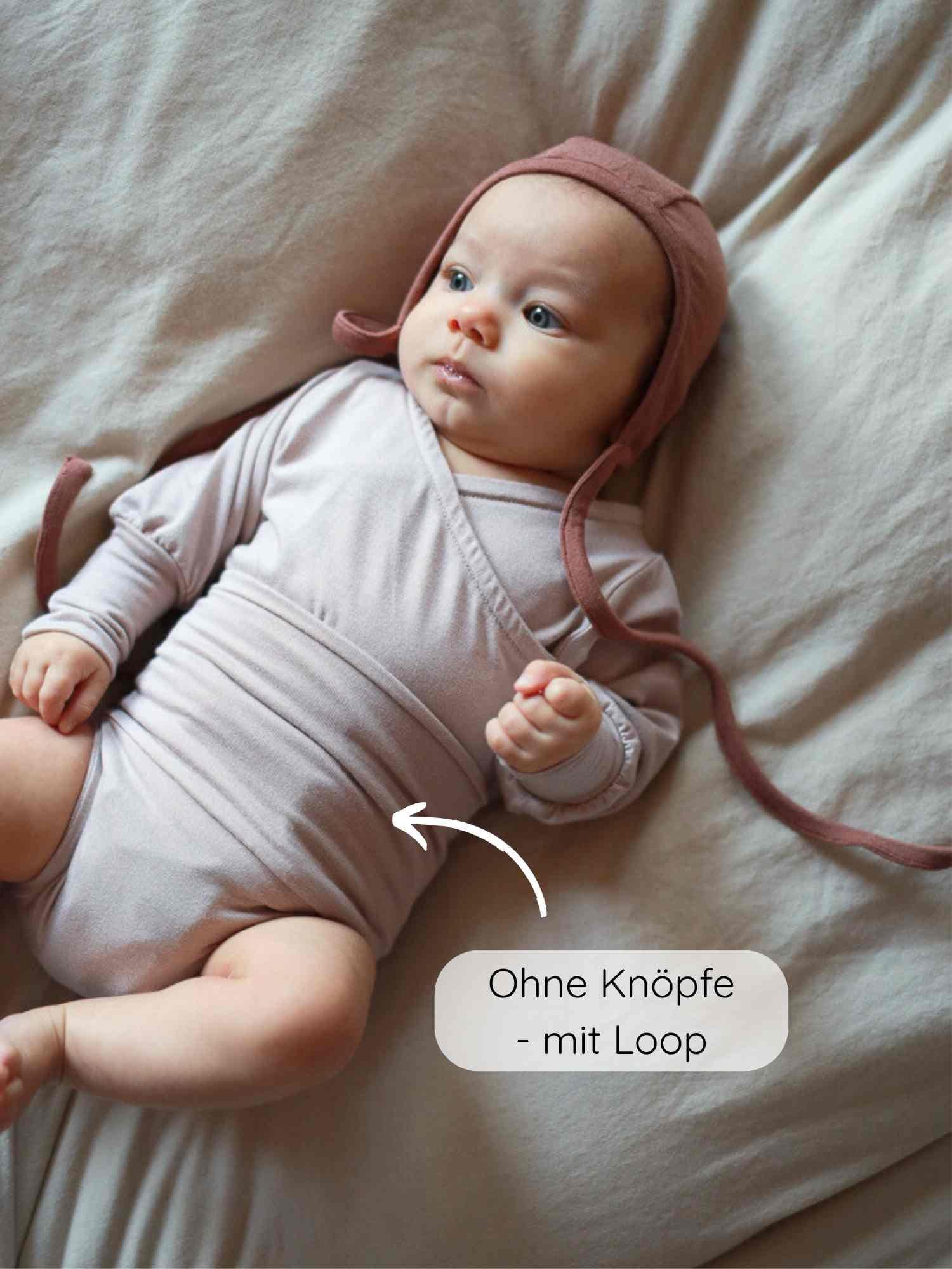 Ein Baby liegt auf einem weichen Laken und trägt ein Loopbody Bambusviskose - Cremepuder, ein knopf- und elastikfreies Wickelbody. Ein Pfeil betont das designpreisgekrönte, wärmende Kleidungsstück.