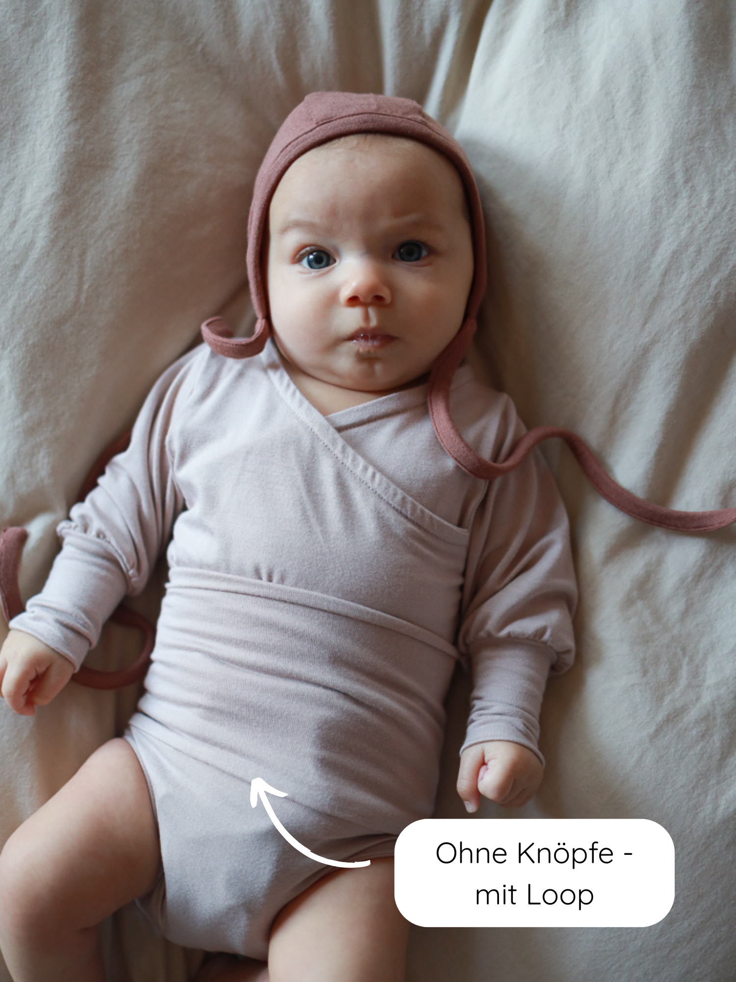 Ein Baby trägt den Loopbody Bambusviskose - Cremepuder, einen staubrosa Wickelbody mit Loop-Verschluss ohne Knöpfe, ergänzt durch eine passende Haube, ideal für bequemes Wickeln und wärmenden Schutz.