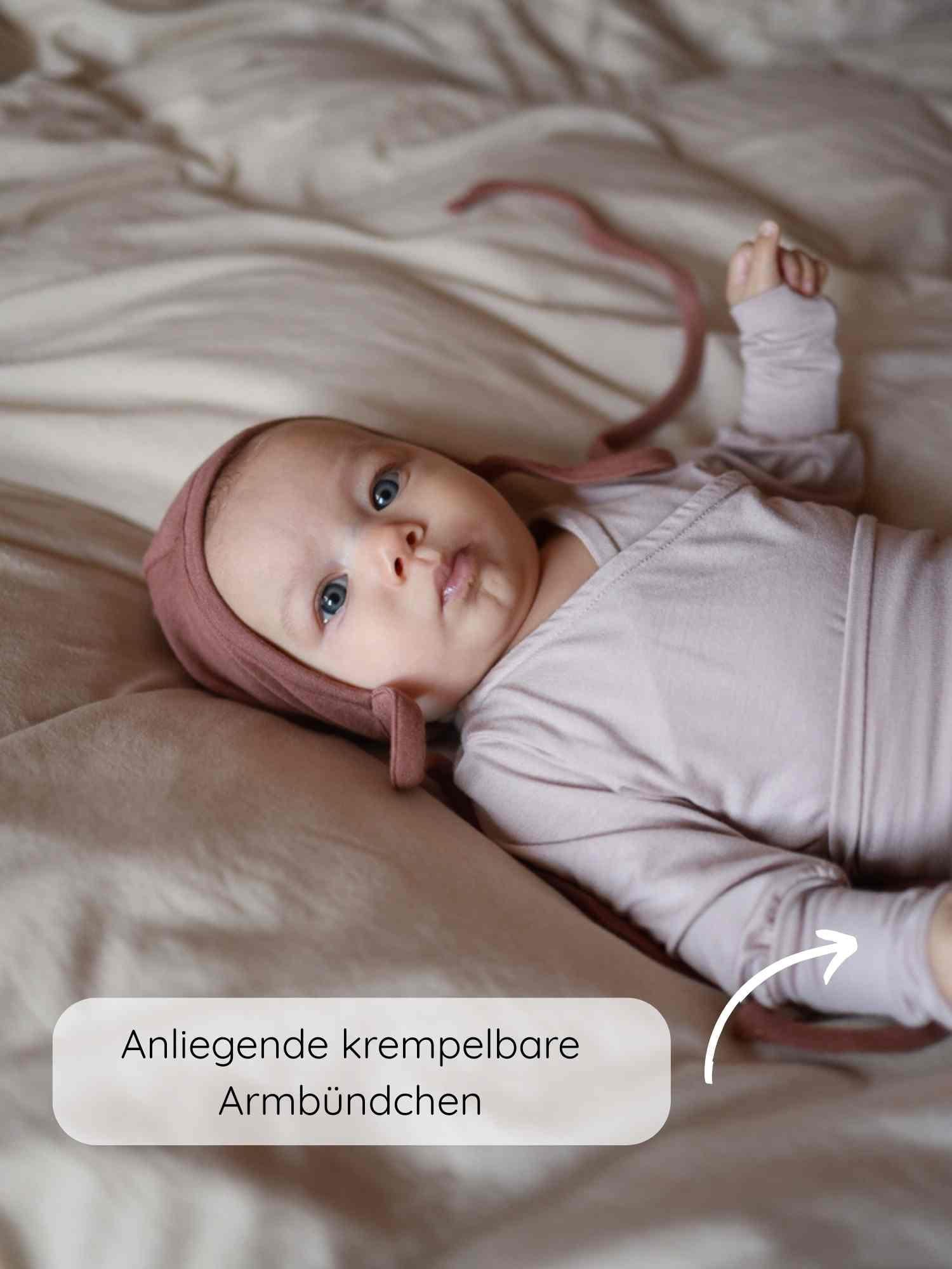 Ein Baby trägt den Loopbody Bambusviskose - Cremepuder, mit anliegenden krempelbaren Armbündchen, auf einer neutralen Bettdecke liegend. Der Onesie hat ein wickelstil Design und eine passende Mütze.