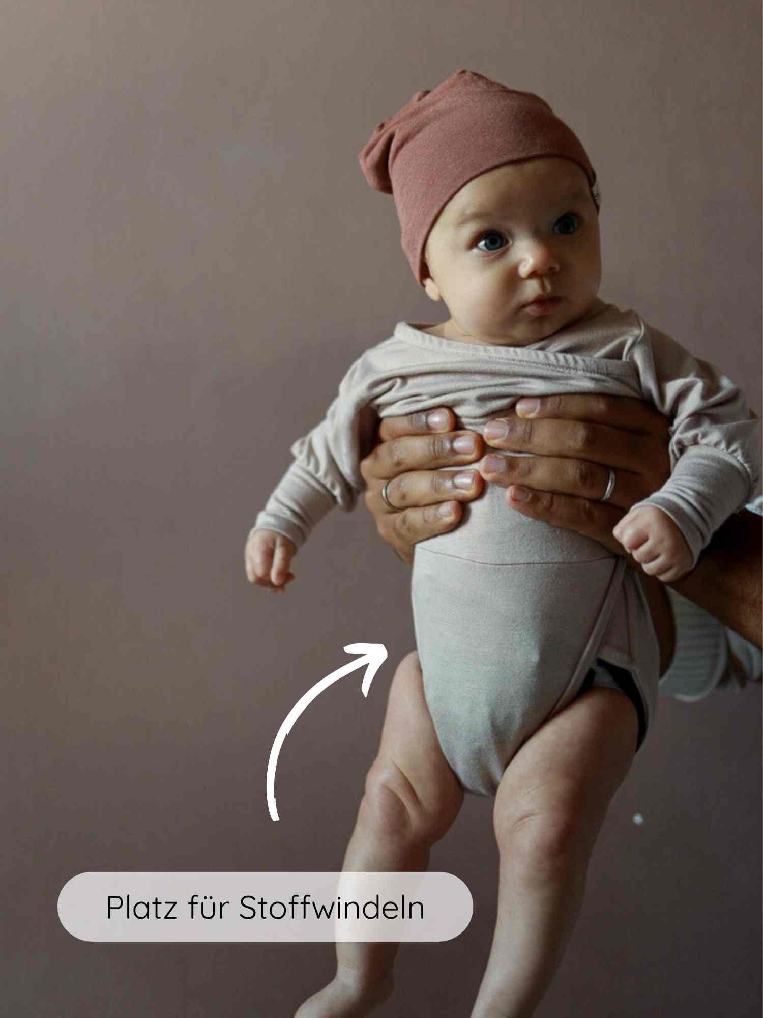 Ein Baby trägt den Loopbody Bambusviskose - Cremepuder, gehalten auf Torsohöhe. Ein Pfeil zeigt auf den Platz für Stoffwindeln, betont durch die Aufschrift „Platz für Stoffwindeln“.