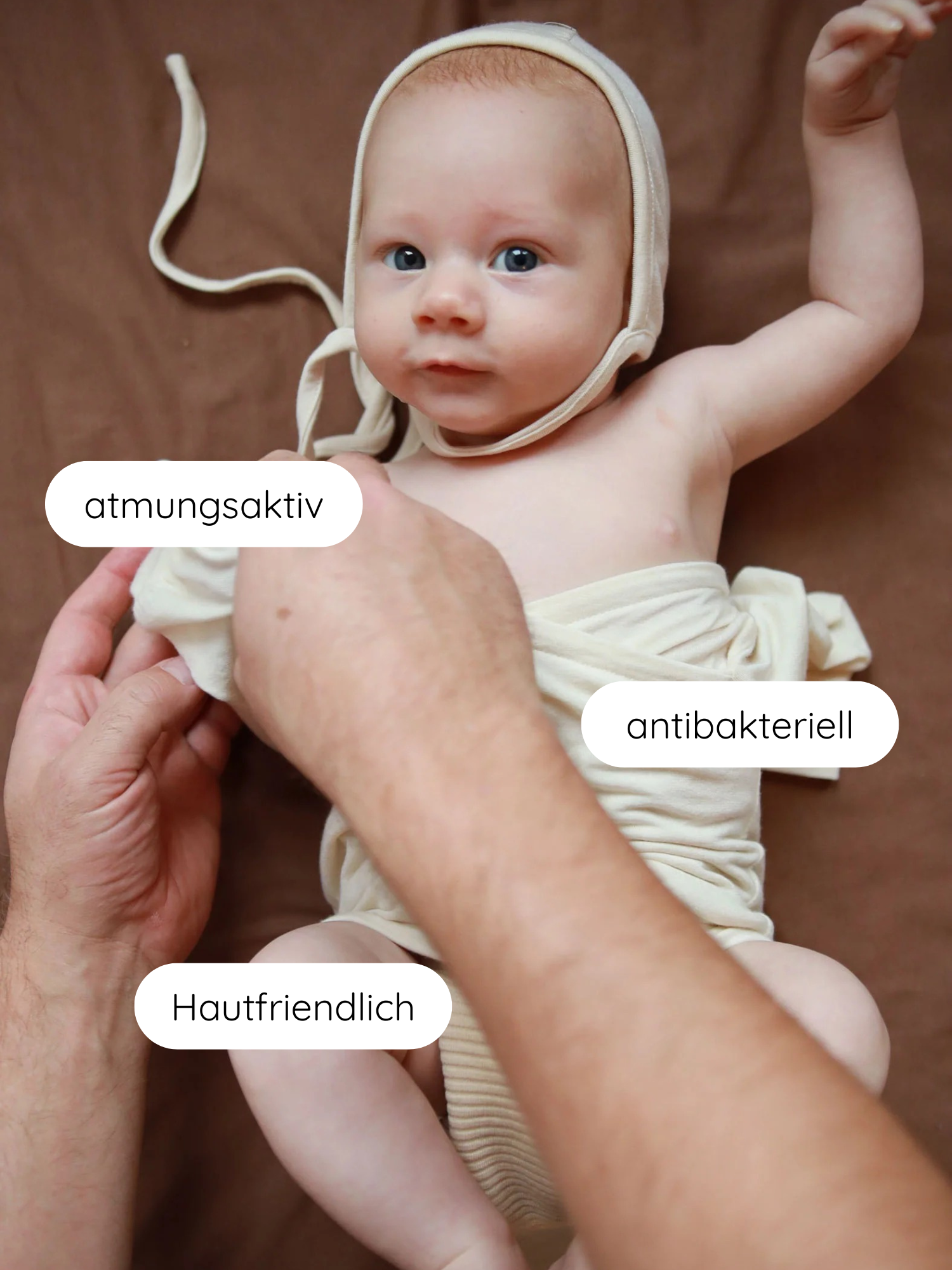 Ein Baby liegt auf einem braunen Untergrund und trägt eine weiche, cremefarbene Haube und ein passendes Wickeltuch aus Bambusviskose. Overlay: atmungsaktiv, antibakteriell, Hautfreundlich. Produkt: Loopbody Bambusviskose - Natur.
