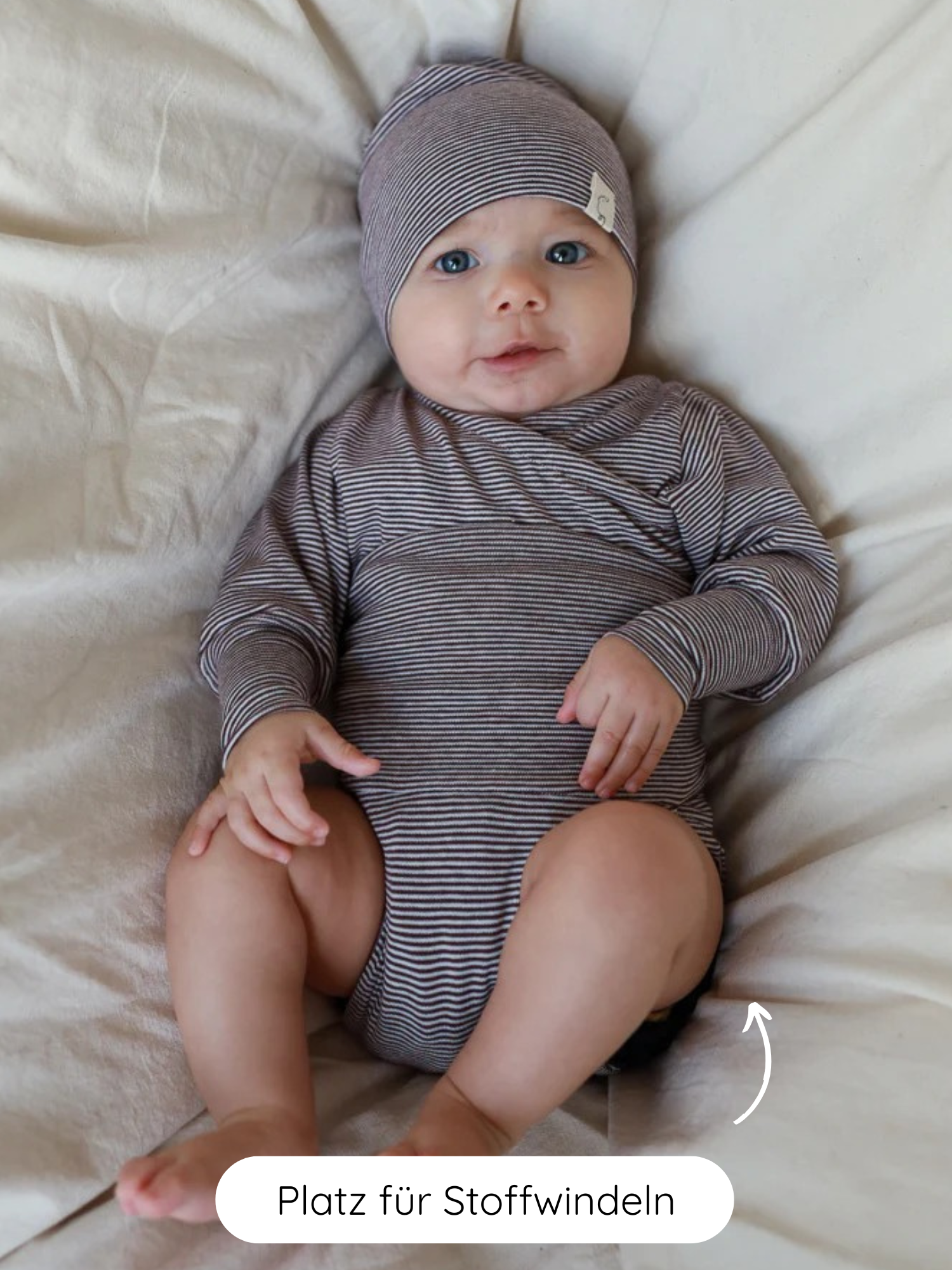 Baby in einem Loopbody Bio-Baumwolle-Ringel Schoko mit passender Mütze, sitzt auf hellem Kissen. Pfeil zeigt Platz für Stoffwindeln, betont die Praktikabilität für Stoffwindelnutzung.