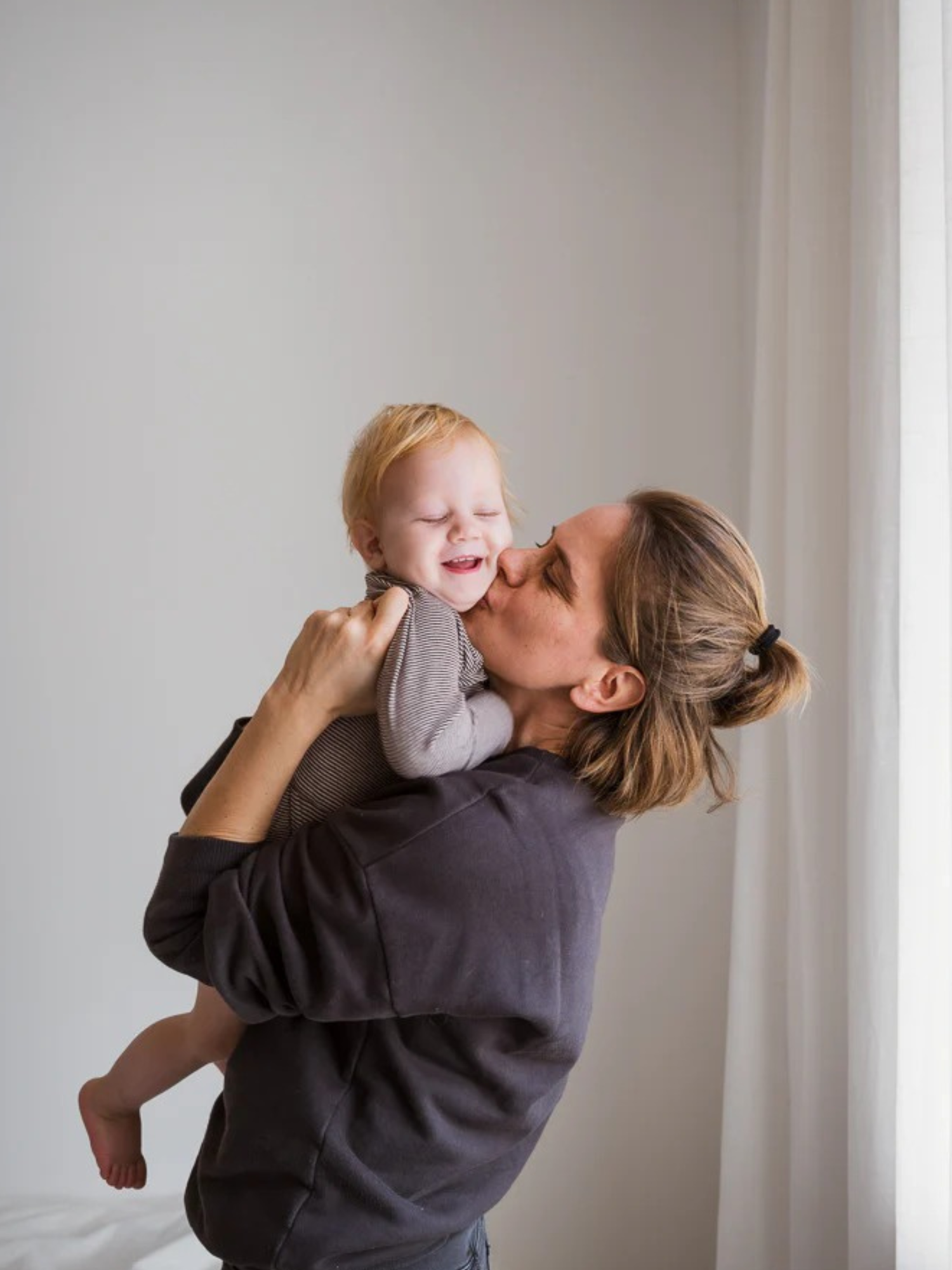 Eine Mutter hebt ihr Baby lächelnd hoch und küsst es. Das Kind trägt den Loopbody Bio-Baumwolle-Ringel Schoko, der bis zu einem Jahr mitwächst, aus weicher Bio-Baumwolle gefertigt.