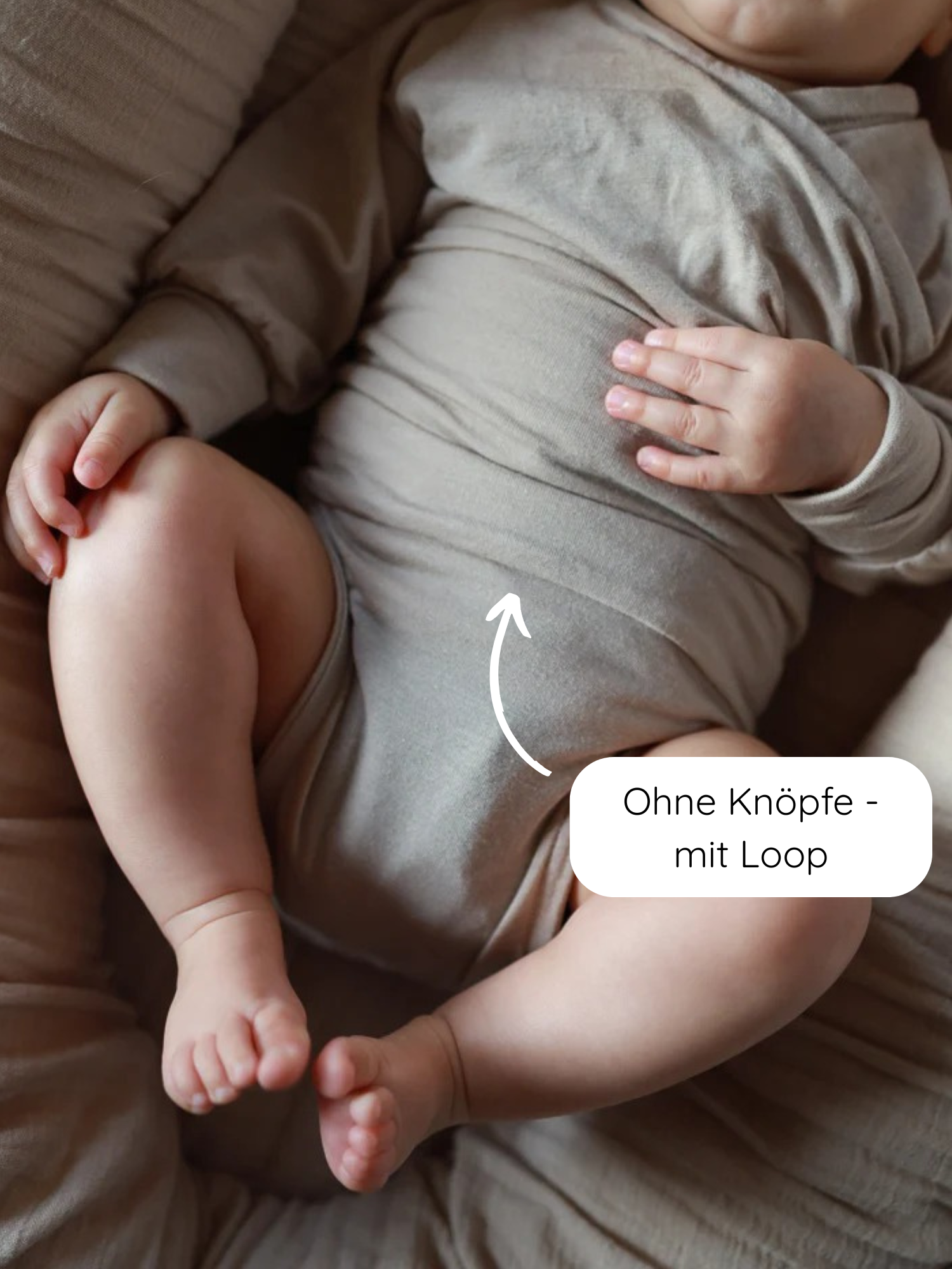 Baby in neutralem, langen Loopbody Kaschmir-Mix - Sand, auf passendem Kissen. Pfeil zeigt auf Mitte des Bodys, Hinweistext: „Ohne Knöpfe – mit Loop“.