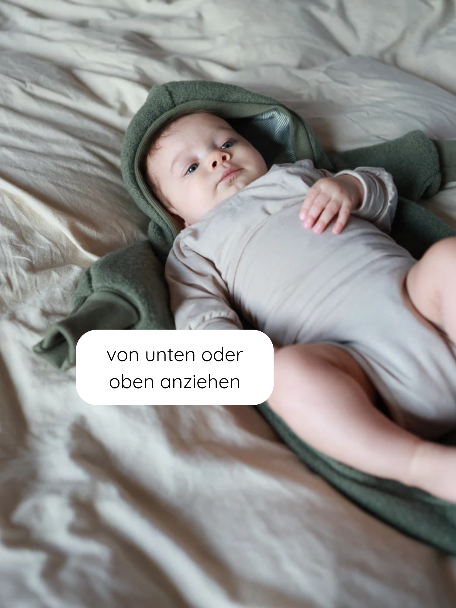 Baby in einem Loopbody Kaschmir-Mix - Sand, liegend auf weicher Decke, teils in grünem Kapuzen-Wrap gehüllt. Text von unten oder oben anziehen im Bild.