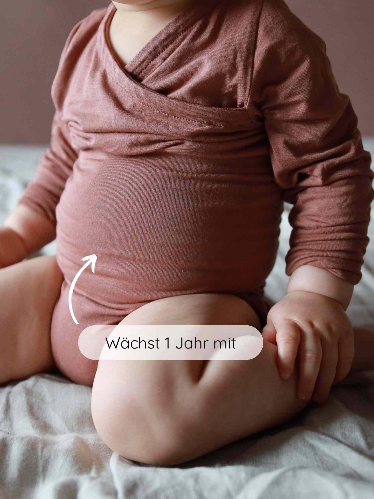 Kleines Kind im Loopbody Kaschmir-Mix - mitwachsender Babybody - Zimt, sitzt bequem auf weichem Untergrund. Weißer Pfeil zeigt auf den Body mit dem Text Wächst 1 Jahr mit.