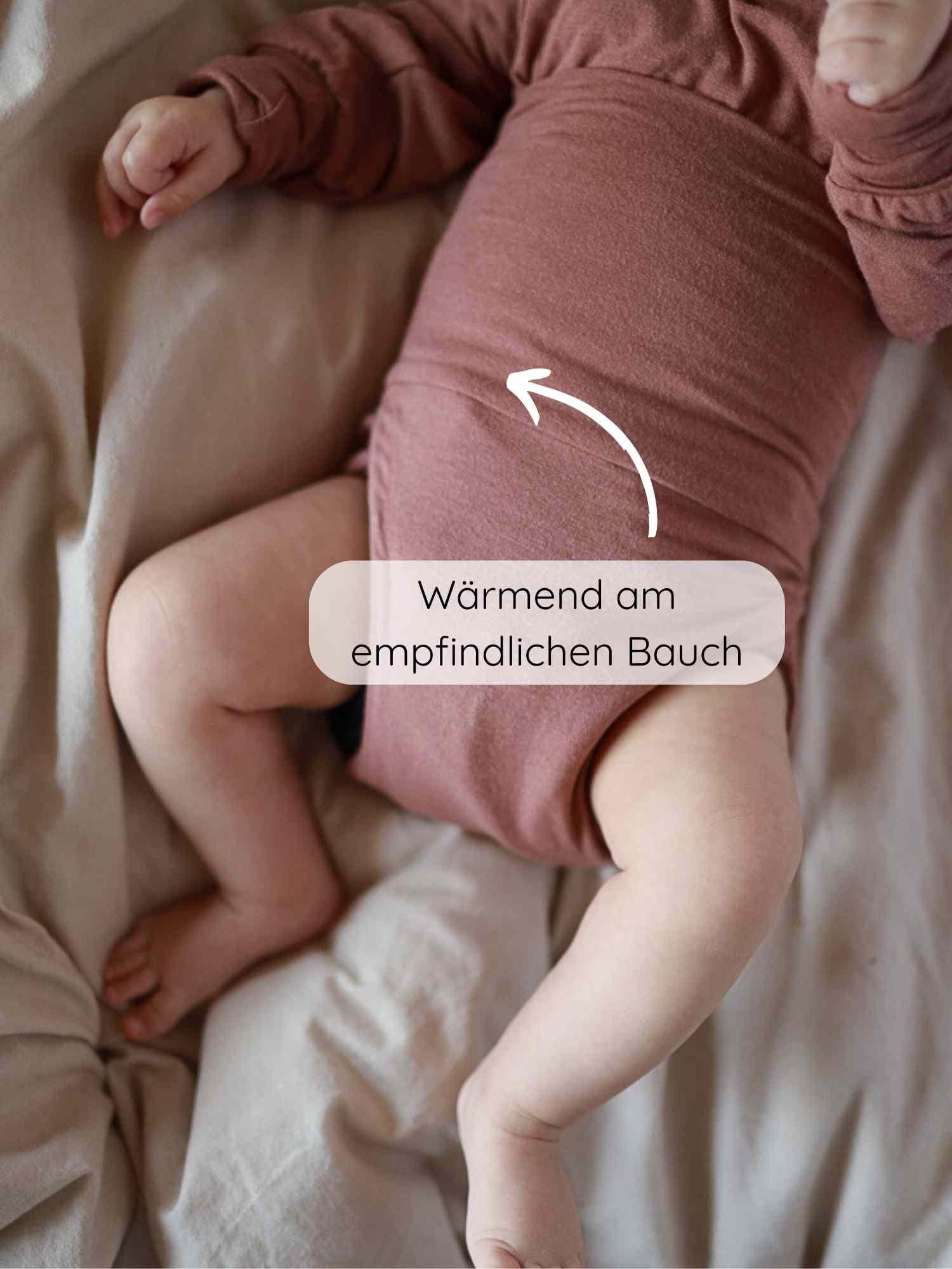 Baby mit Loopbody Kaschmir-Mix - mitwachsender Babybody - Zimt, auf einem Bett liegend. Weißer Pfeil zeigt auf den Bauch mit dem eingebauten Wärmer, Text: Wärmend am empfindlichen Bauch.