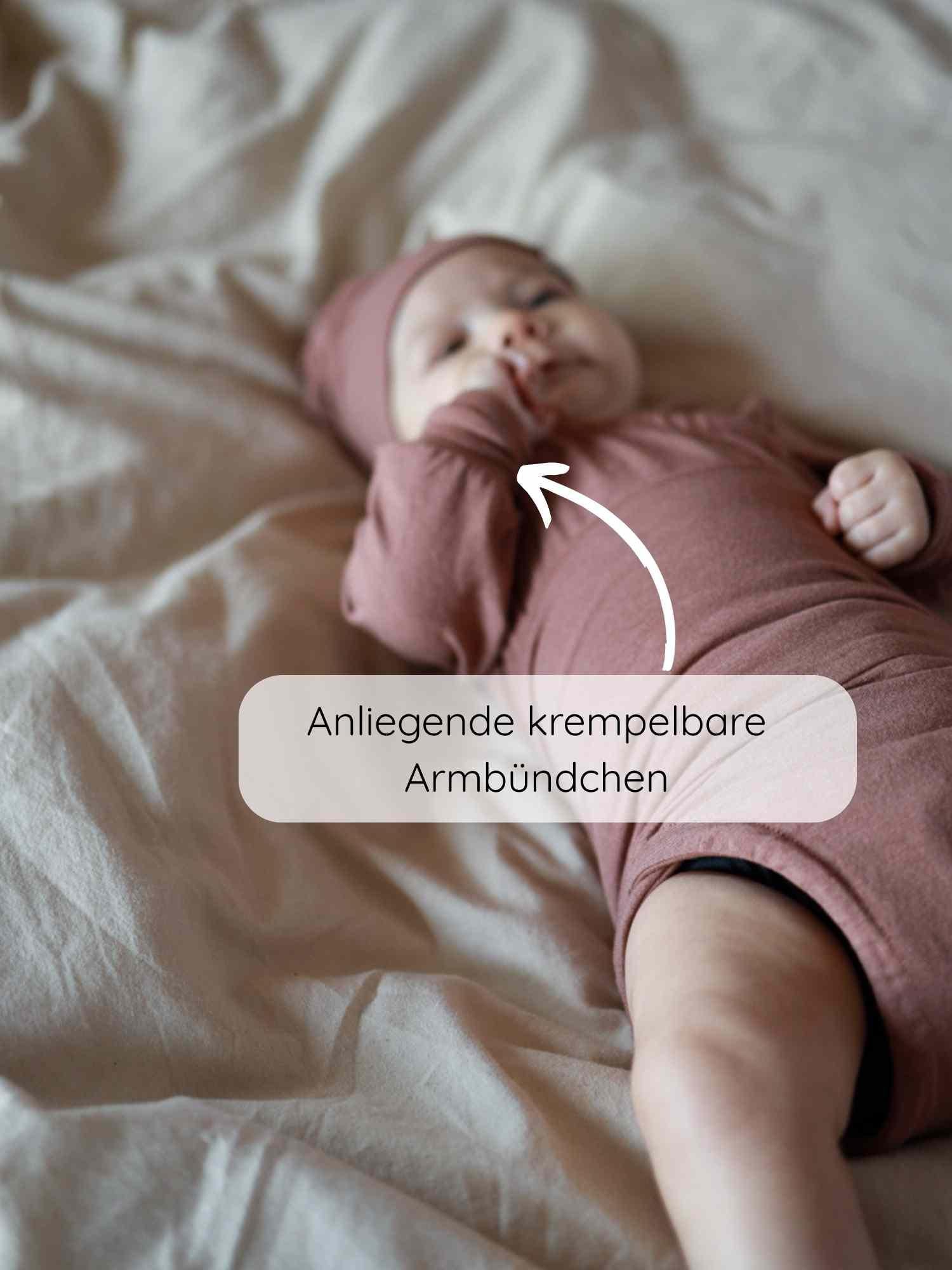 Loopbody Kaschmir-Mix - mitwachsender Babybody - Zimt: Baby in braun-rosa Langarmbody mit passender Mütze, weiße Pfeile zeigen auf krempelbare Armbündchen, weiches, bequemes Material für lange Tragbarkeit.