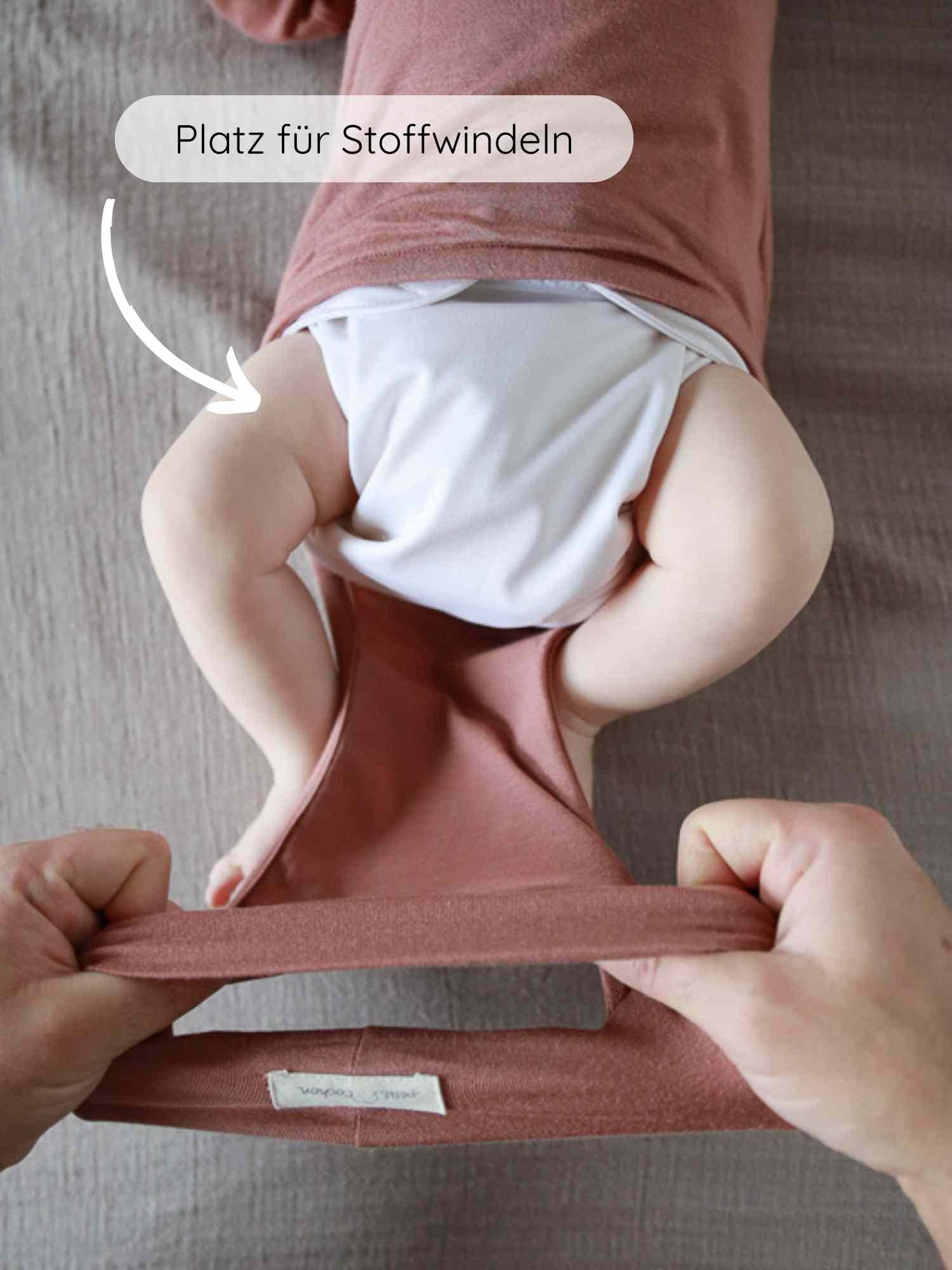 Loopbody Kaschmir-Mix - mitwachsender Babybody - Zimt: Ein Baby trägt einen weißen Stoffwindel unter einem rostrosa Wickelbody. Zwei Hände dehnen den Saum, um Platz für Windeln zu zeigen.