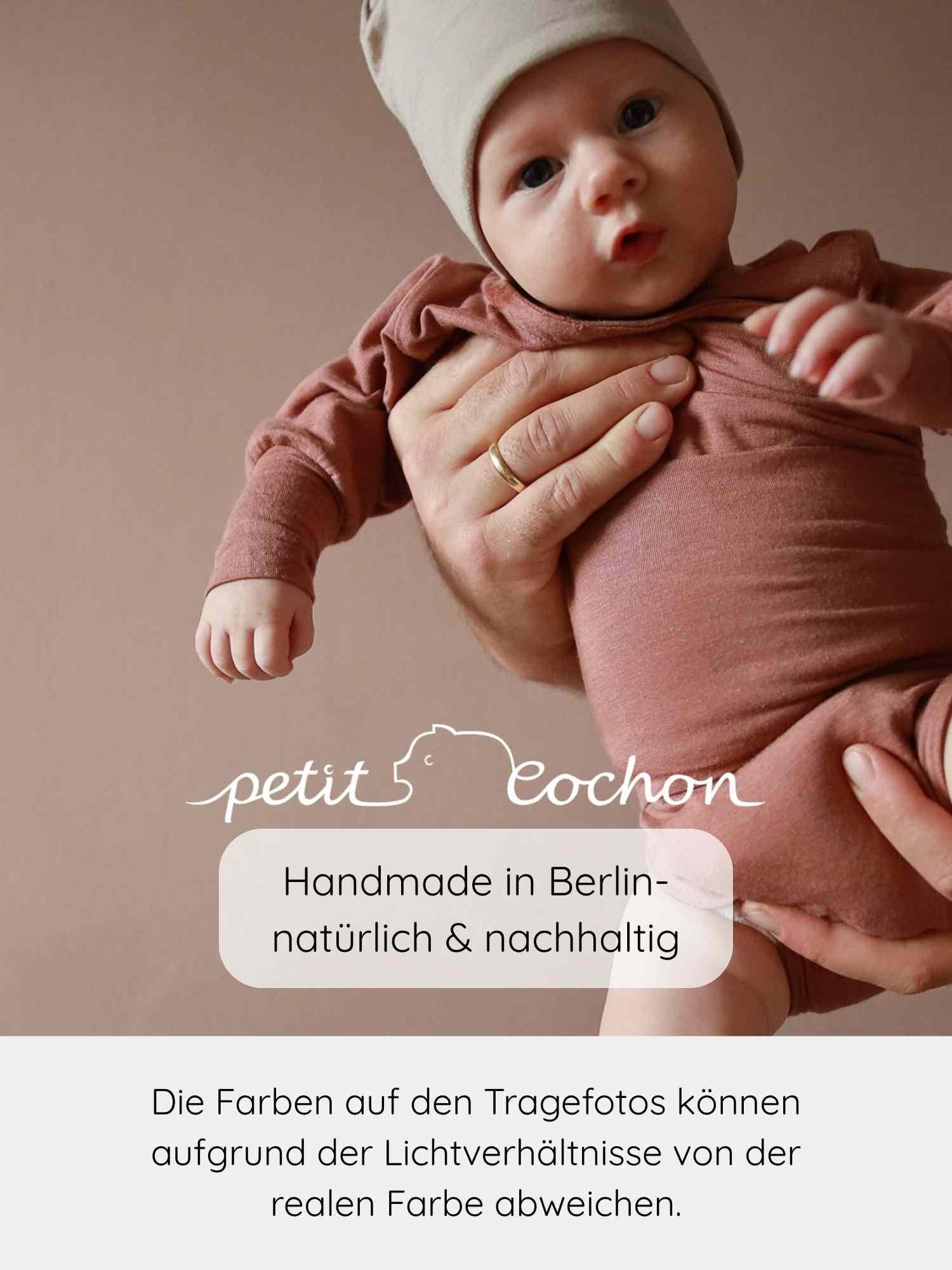 Baby in rostbraunem Loopbody Kaschmir-Mix - mitwachsender Babybody - Zimt, gehalten von einer erwachsenen Hand. Beanie passend zum Body. Überlagert mit petit cochon Logo und Berliner Handwerksqualität.