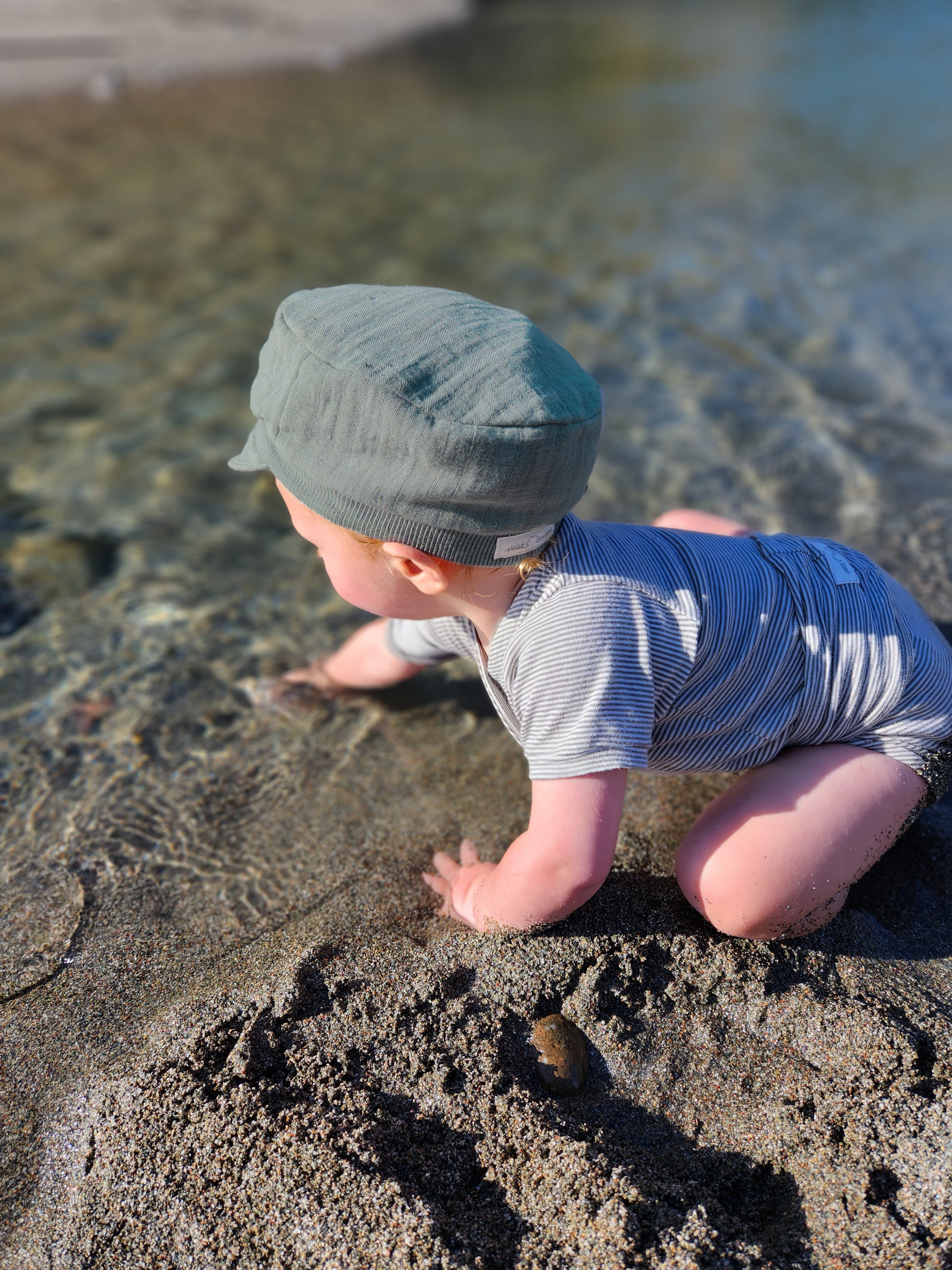 Ein Baby spielt am Strand und trägt die Michelmütze Bio-Musselin - Taube, eine leichte Sommermütze mit elastischem Bündchen aus nachhaltigem Musselin.