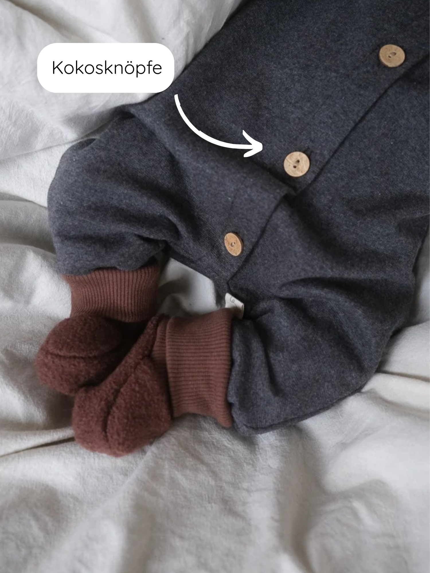 Mini Overall Feinstrick - Ruß Meliert auf weißem Untergrund, zeigt Kokosknöpfe und braune Bündchen. Der Overall wird von weichen, braunen Teddy-Fleece-Booties ergänzt und ist aus meliertem Feinstrick-Jersey gefertigt.